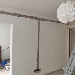 Rafał Klimczyk Elektryka - Przygotowanie bruzd pod instalację elektryczną w ścianie. Widoczne kable i narzędzia. W tle biała lampa i drabina. Etap prac przed tynkowaniem.