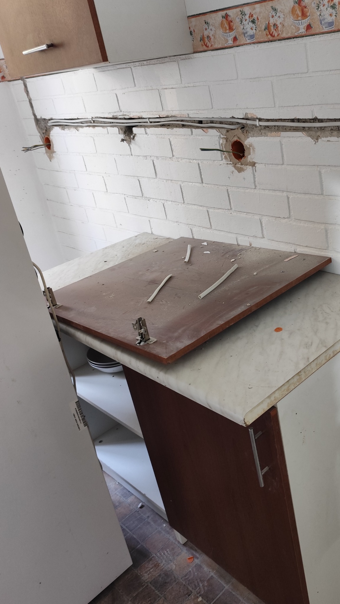 Biała ściana z cegły z przewodami elektrycznymi w bruzdach, częściowo zdemontowane szafki kuchenne z brązowym blatem i elementami instalacji elektrycznej.