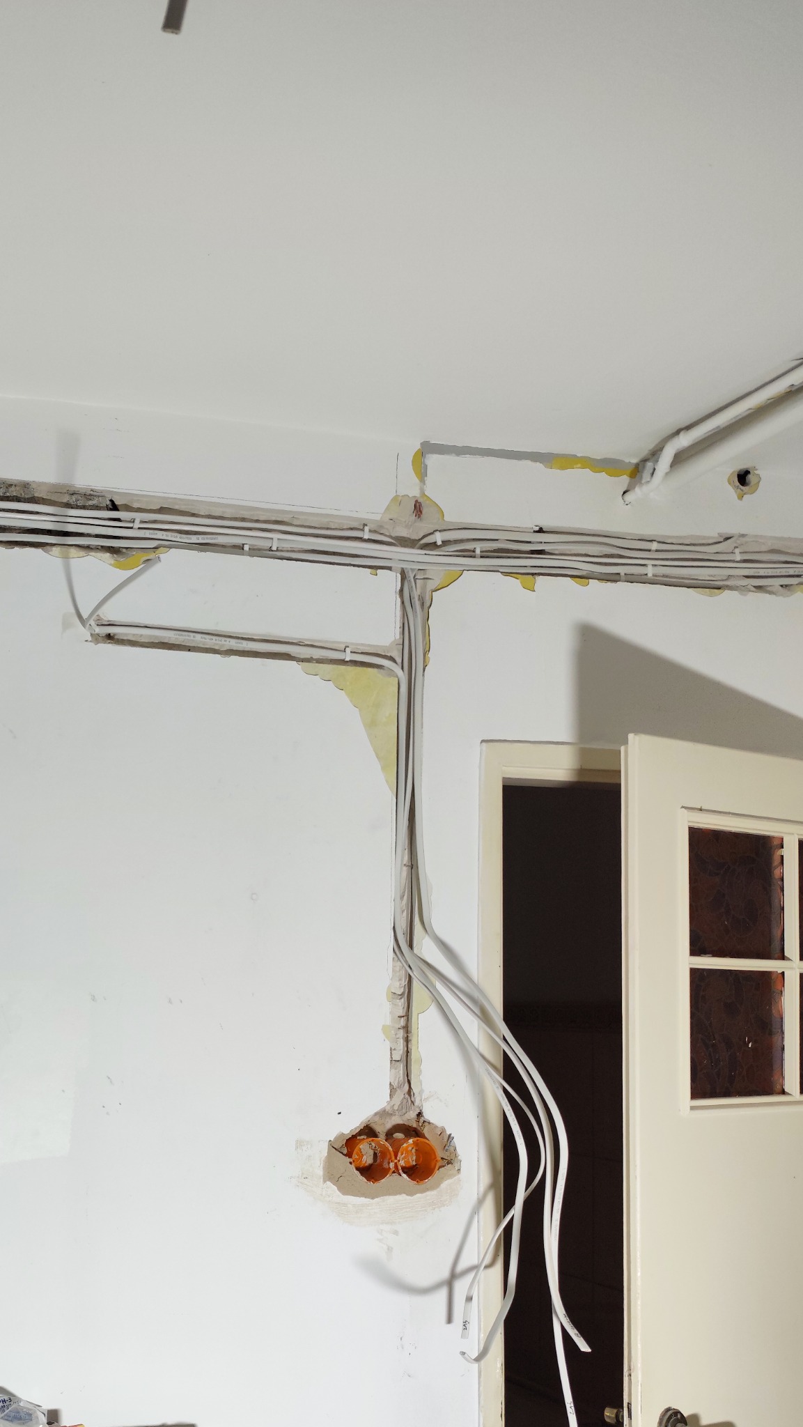 Biała ściana z widoczną instalacją elektryczną w trakcie montażu. Kable prowadzone w listwach i puszkach, ściana częściowo obita. Widoczne fragmenty drzwi.