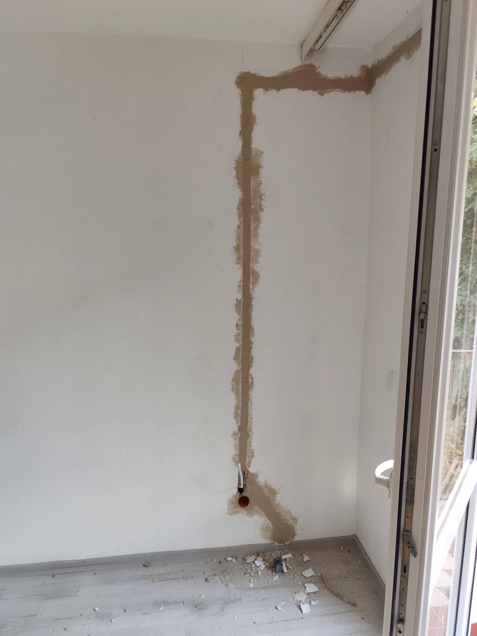 Biała ściana z wyżłobionymi bruzdami pod instalację elektryczną, widoczne kable w peszlu. Fragment okna po prawej stronie. Podłoga z panelami.