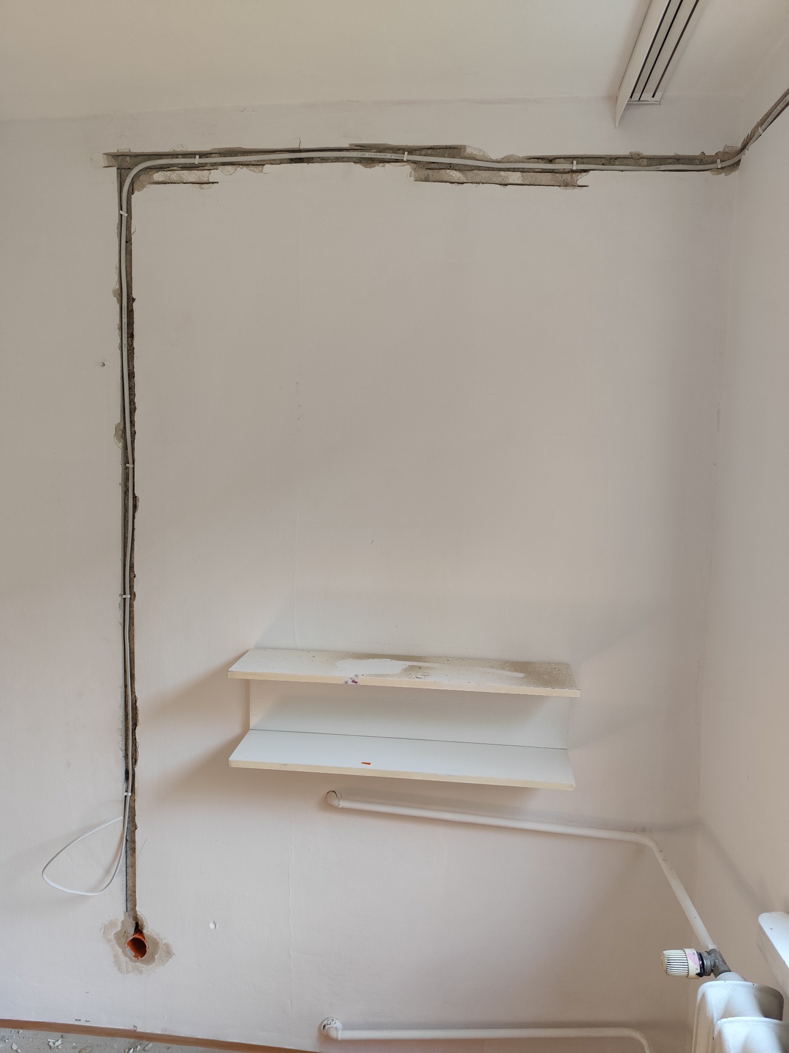 Biała ściana z wyżłobieniami pod kable elektryczne, częściowo zamaskowane gipsem. Widoczne przewody i rurka instalacyjna w ścianie. Dwie półki na ścianie.