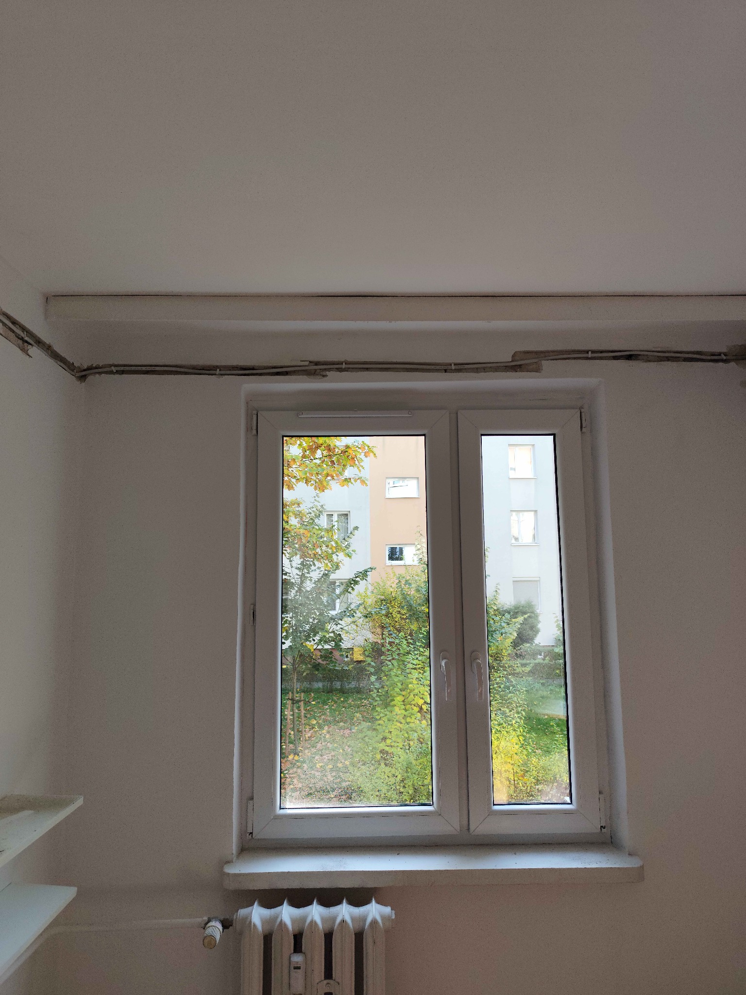 Białe okno PCV z widokiem na zieleń, instalacja elektryczna pod sufitem, grzejnik żeberkowy pod parapetem. Widoczne kable w listwach.