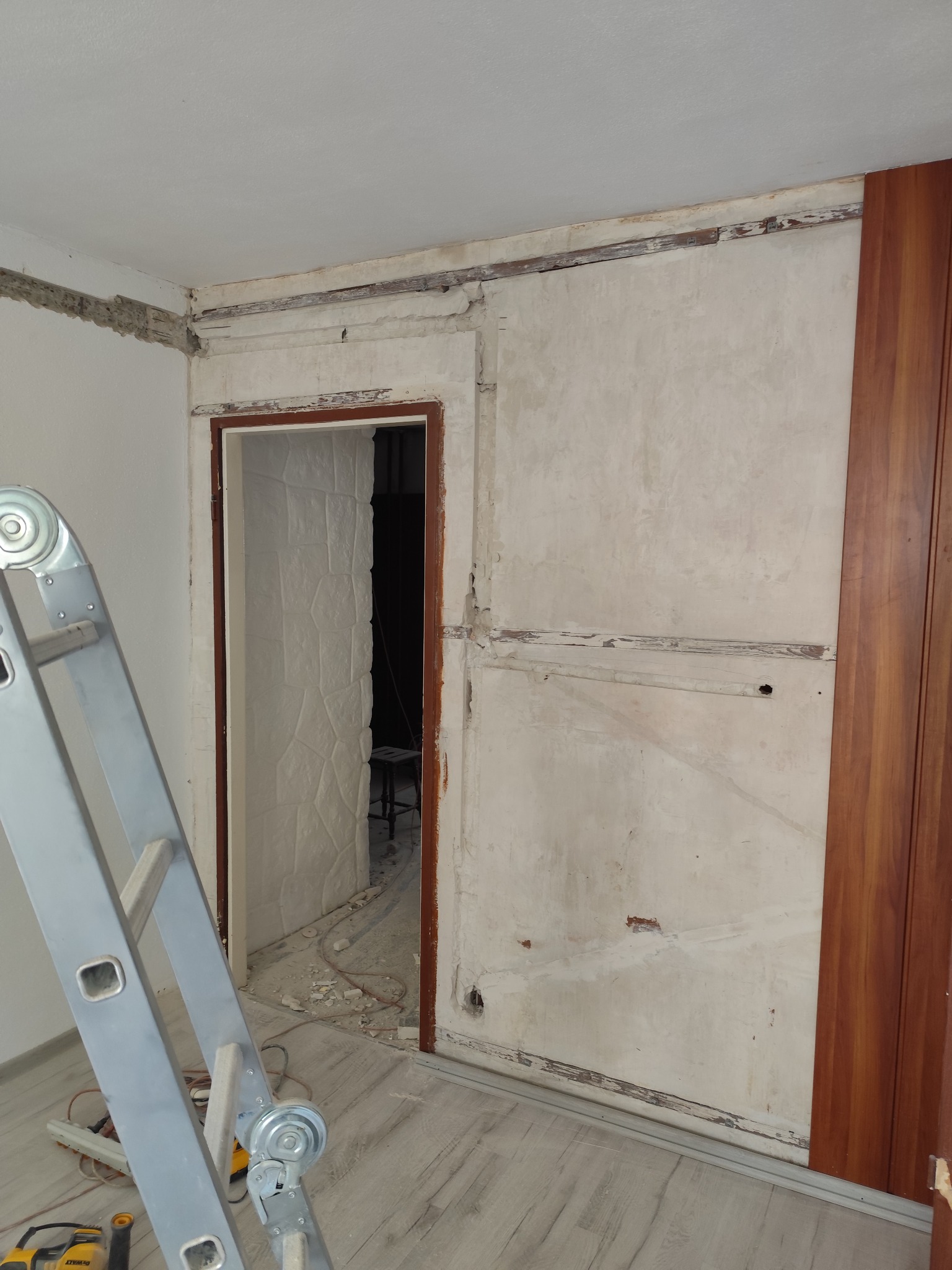 Remont pokoju: ściana w trakcie przygotowania, widoczne ślady po instalacjach, drabina oparta o ścianę, fragment szafy przesuwnej. Prace wykończeniowe w toku.