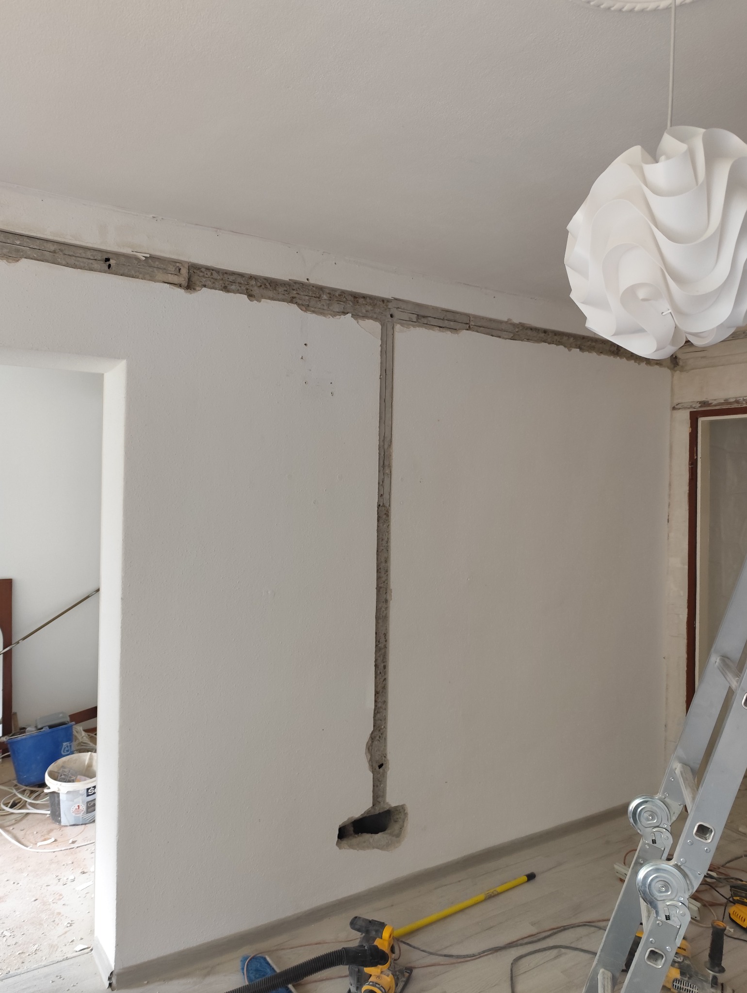 Przygotowanie bruzd pod instalację elektryczną w ścianie. Widoczne kable i narzędzia. W tle biała lampa i drabina. Etap prac przed tynkowaniem.