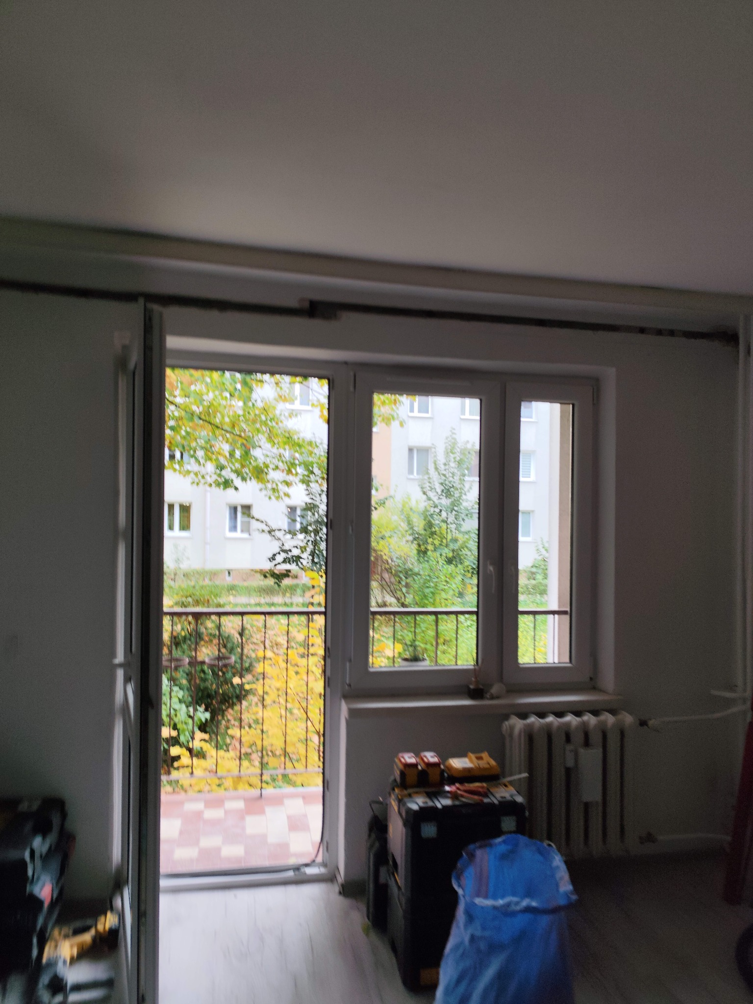 Wnętrze pokoju z oknem balkonowym, widoczne narzędzia i skrzynka elektryczna, sugerujące prace instalacyjne. Jesienne drzewa za oknem dodają koloru.