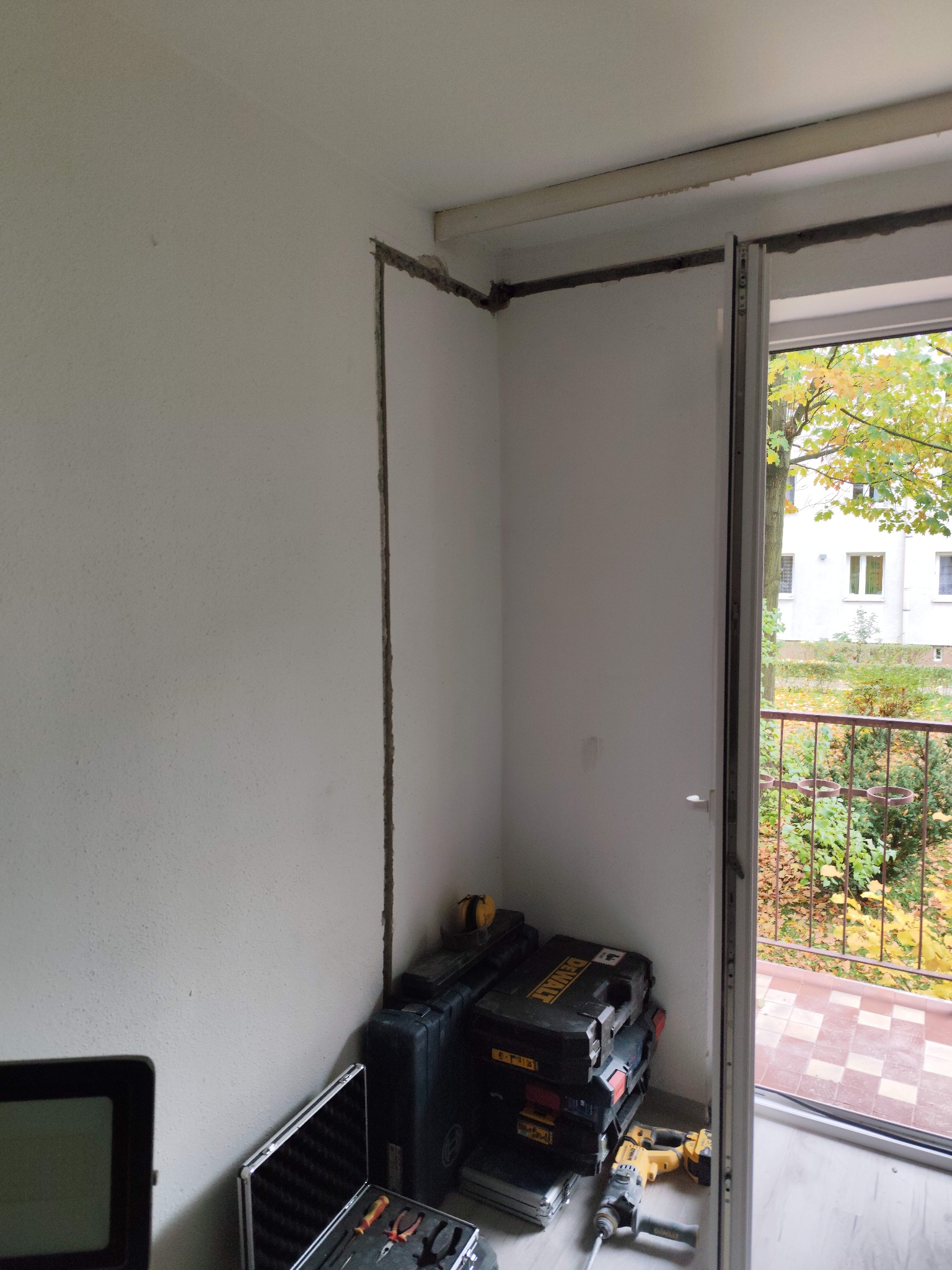 Przygotowanie pod instalację elektryczną: wyżłobione bruzdy w ścianie, widoczne narzędzia i skrzynki narzędziowe DeWalt. Balkon z widokiem na zieleń w tle.