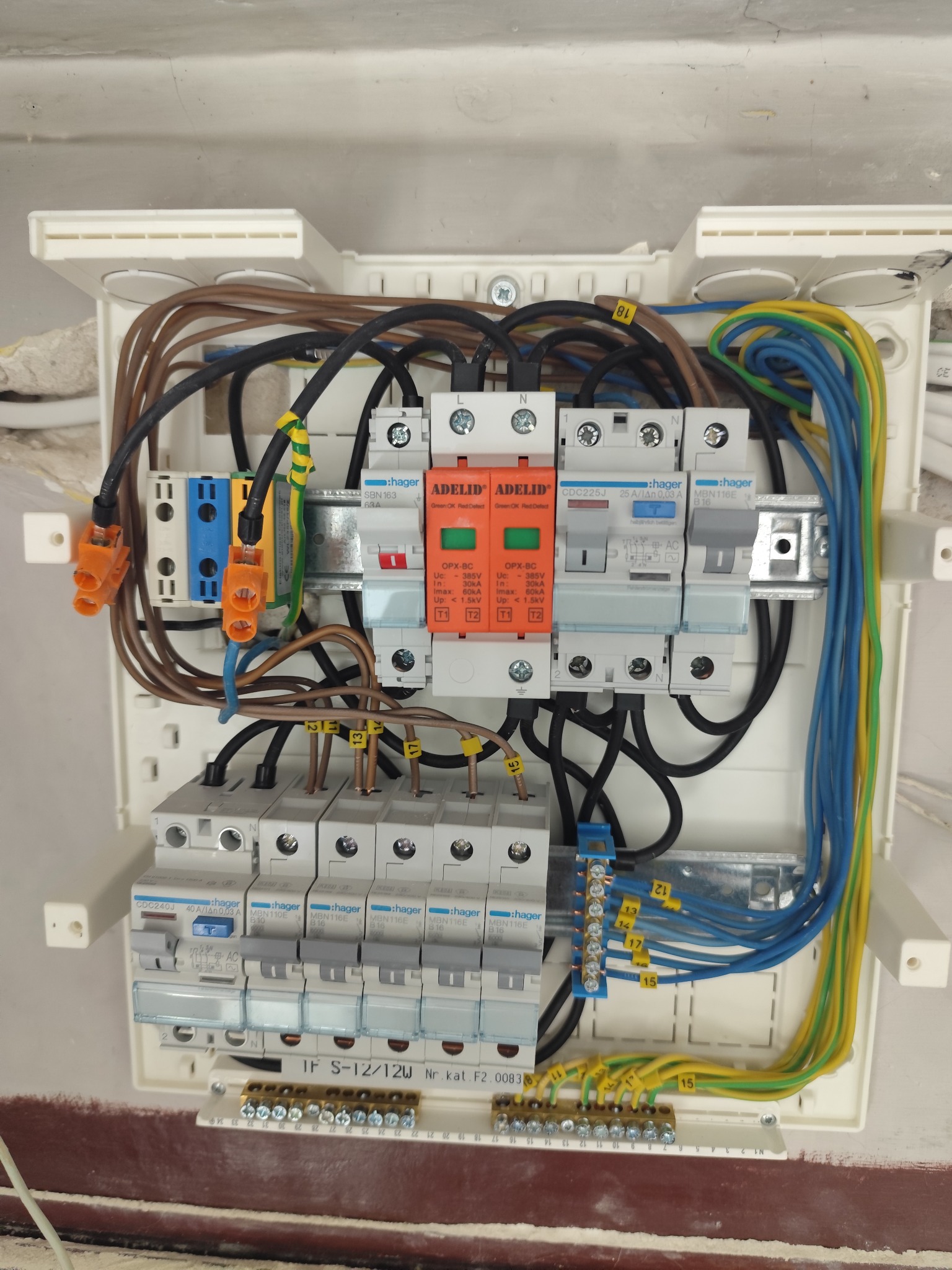 Rozdzielnica elektryczna w białej obudowie z widocznymi bezpiecznikami, przewodami oznaczonymi numerami i kolorami, oraz elementami marki Hager i Adelid.