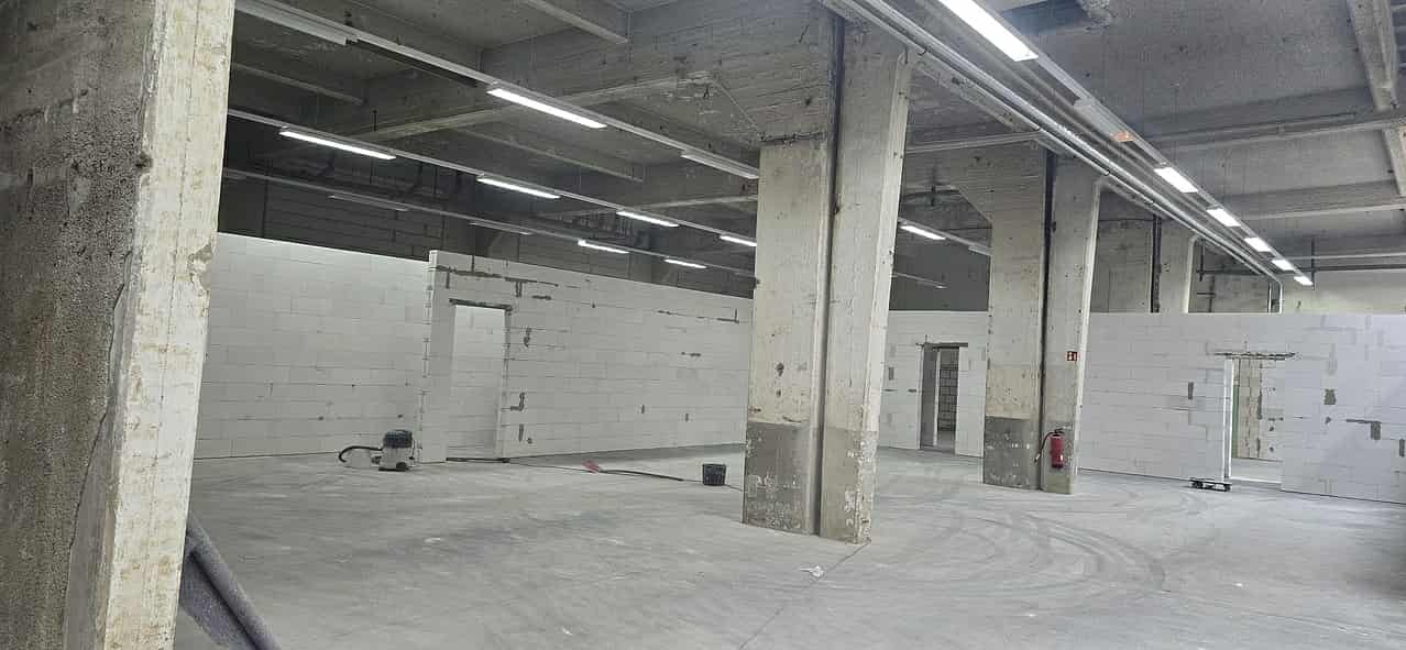 Wnętrze hali w trakcie budowy ścian działowych z betonu komórkowego. Widoczne surowe słupy i instalacja oświetleniowa pod stropem. Na podłodze widoczny odkurzacz.