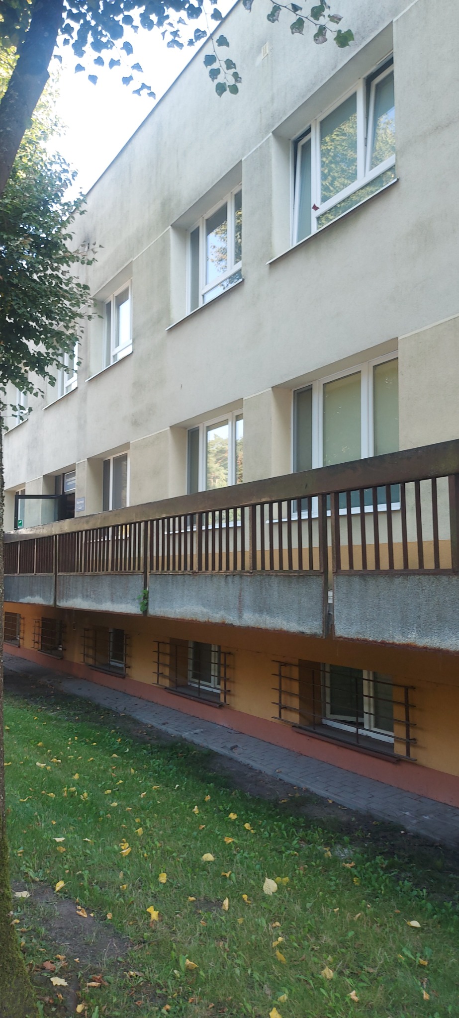 Budynek z drewnianą balustradą i metalowymi kratami w oknach piwnicznych, widok z zewnątrz, trawnik z opadłymi liśćmi, drzewo po lewej stronie.