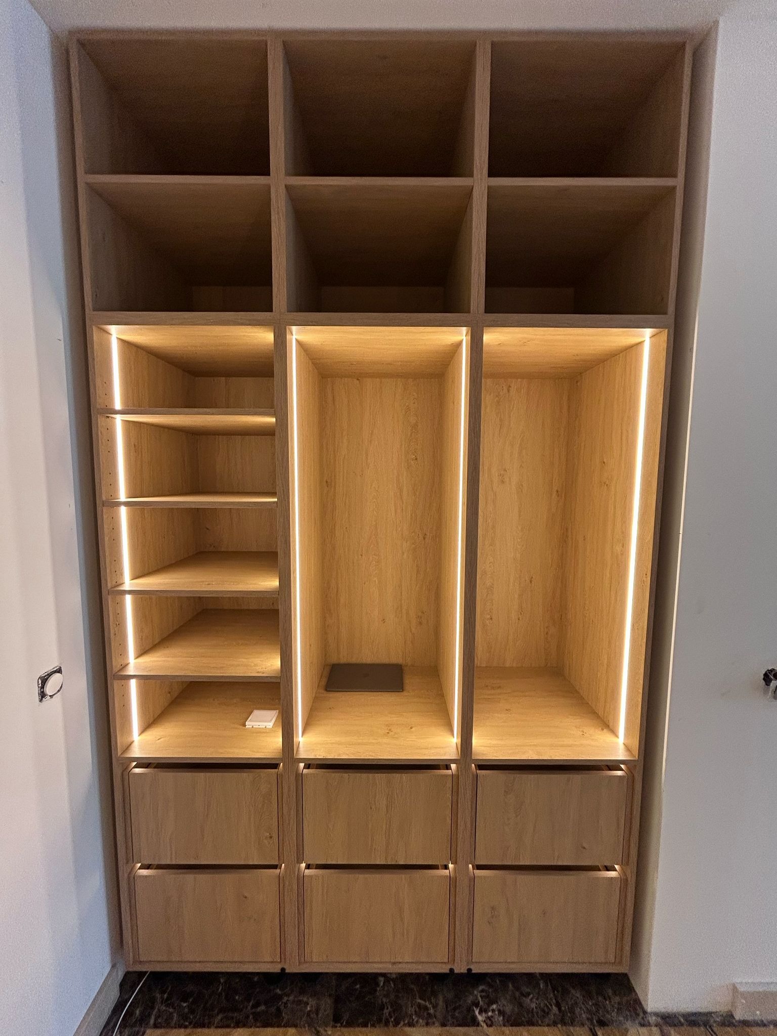 Drewniana szafa wnękowa z oświetleniem LED, widoczne półki i szuflady. Na półce leży biały przedmiot. Marmurowa podłoga.