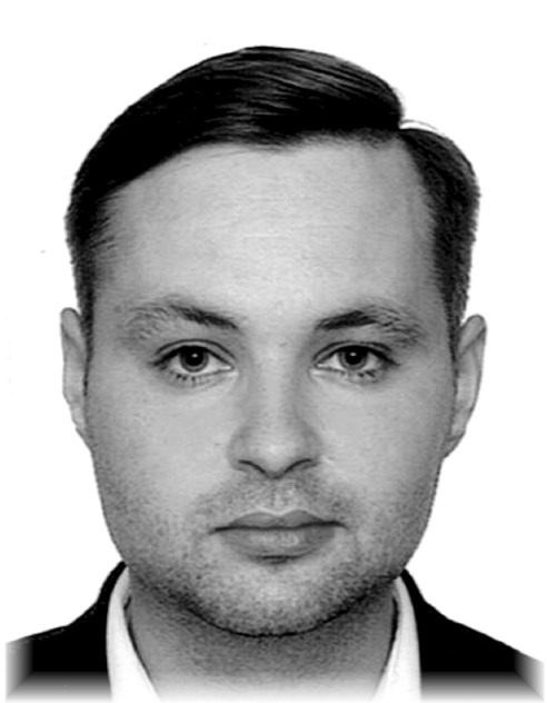 Portret mężczyzny w czarno-białej tonacji, z krótkimi włosami i zarostem, ubrany w ciemny garnitur i jasną koszulę. Ujęcie od przodu, z delikatnym cieniowaniem.