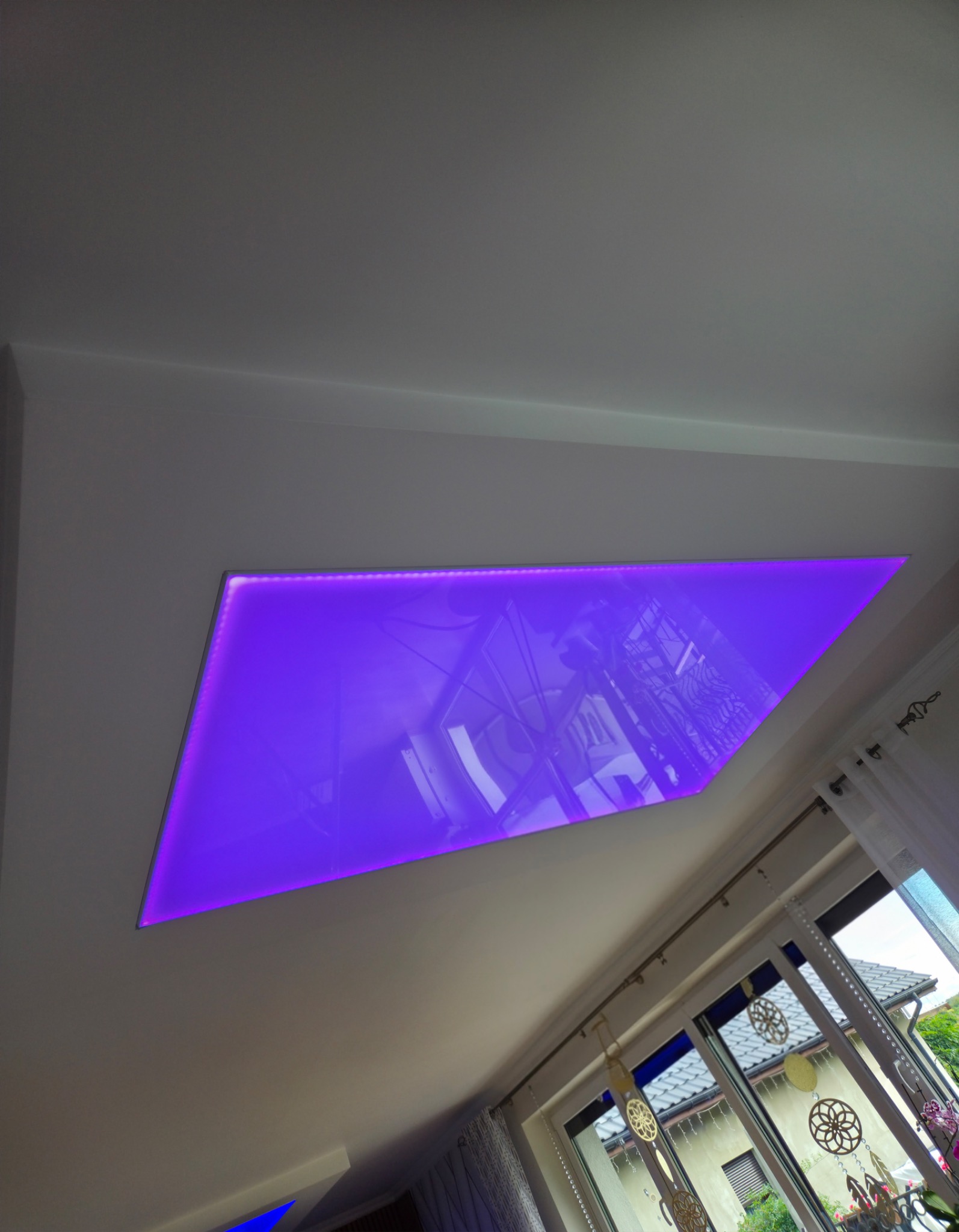 Nowoczesny sufit podwieszany z podświetleniem LED w kolorze fioletowym, widok z dołu. W tle okno z dekoracjami.