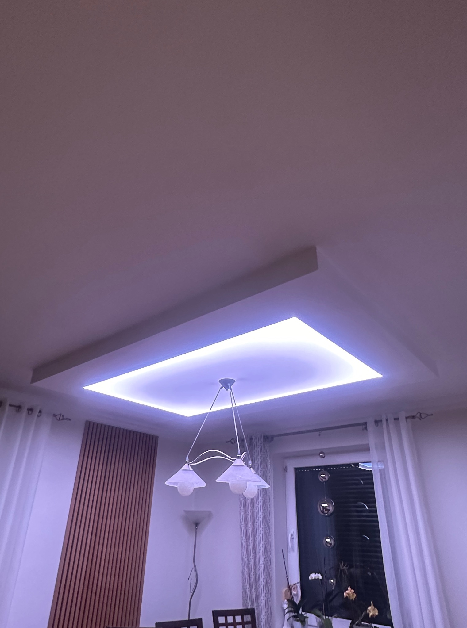 Biały sufit podwieszany z oświetleniem LED w kształcie kwadratu, wisząca lampa, okno z roletami i zasłonami, oraz dekoracyjne panele ścienne.