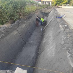AQUAFORM SPÓŁKA Z OGRANICZONĄ ODPOWIEDZIALNOŚCIĄ - Betonowy rów melioracyjny w trakcie budowy, dwóch robotników w środku, widok z góry, elementy infrastruktury drogowej w tle.