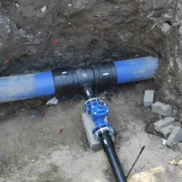 Połączenie niebieskiego rurociągu z zaworem w wykopie ziemnym. Widoczne złącza, rury z tworzywa i fragmenty gruzu na dnie wykopu. Instalacja wodociągowa w trakcie montażu.