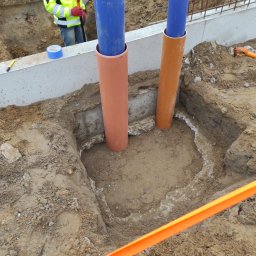 AQUAFORM SPÓŁKA Z OGRANICZONĄ ODPOWIEDZIALNOŚCIĄ - Wykop pod instalacje w gruncie we Wrocławiu, zabezpieczony pomarańczową taśmą. Widoczne wystające rury w osłonach. Pracownik w tle.