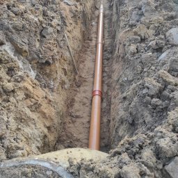 AQUAFORM SPÓŁKA Z OGRANICZONĄ ODPOWIEDZIALNOŚCIĄ - Brązowa rura kanalizacyjna z czerwonym złączem ułożona w wykopie ziemnym. Widoczne ślady pracy ziemnej i fragment betonowego kręgu.