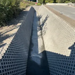 Długi wykop wyłożony ażurowymi betonowymi płytami, prawdopodobnie pod instalacje. Widoczny fragment terenu z zielenią i element infrastruktury drogowej w tle.