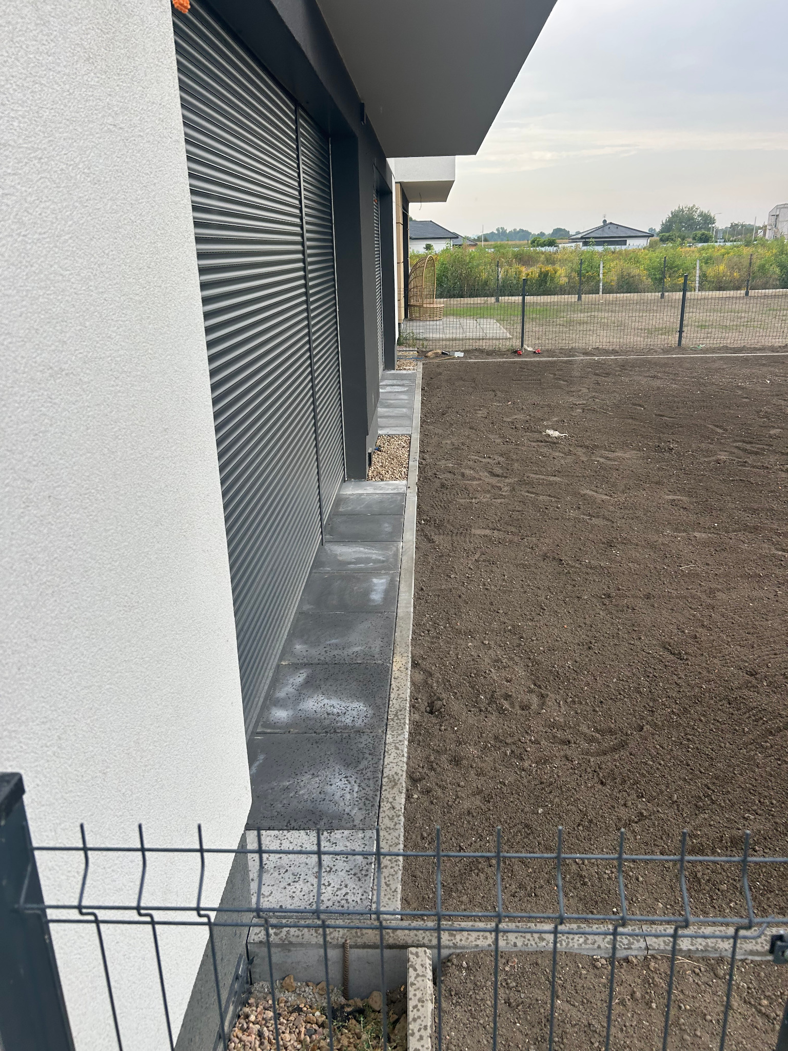 Przygotowany teren pod trawnik przy nowoczesnym domu z szarą elewacją i roletami zewnętrznymi. Widoczna siatka ogrodzeniowa i ułożone płyty chodnikowe.