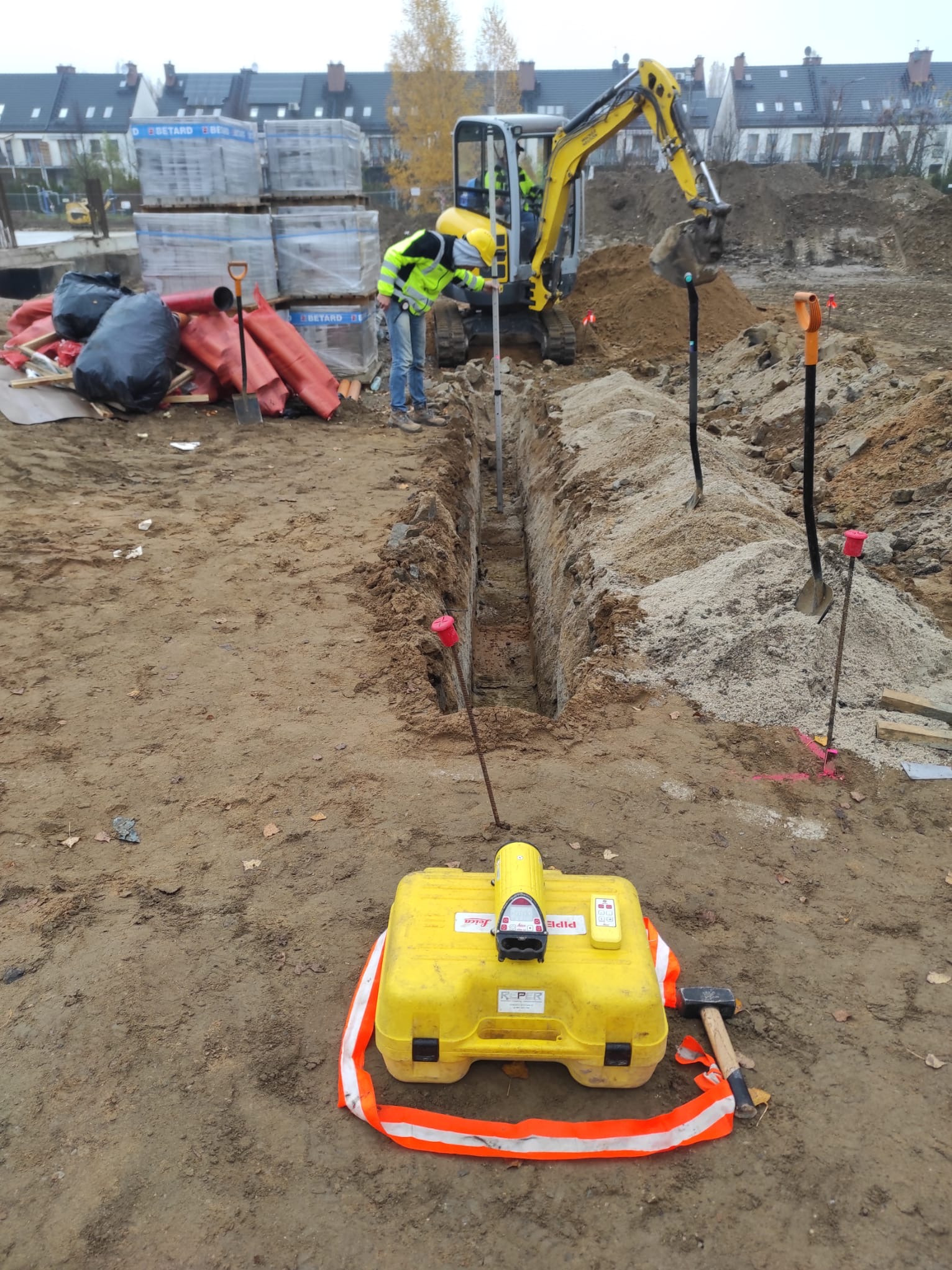 Wykop wąskoprzestrzenny pod instalacje, z żółtym detektorem rur na pierwszym planie. Mini koparka w tle, pracownik w odblaskowej kamizelce przy wykopie.