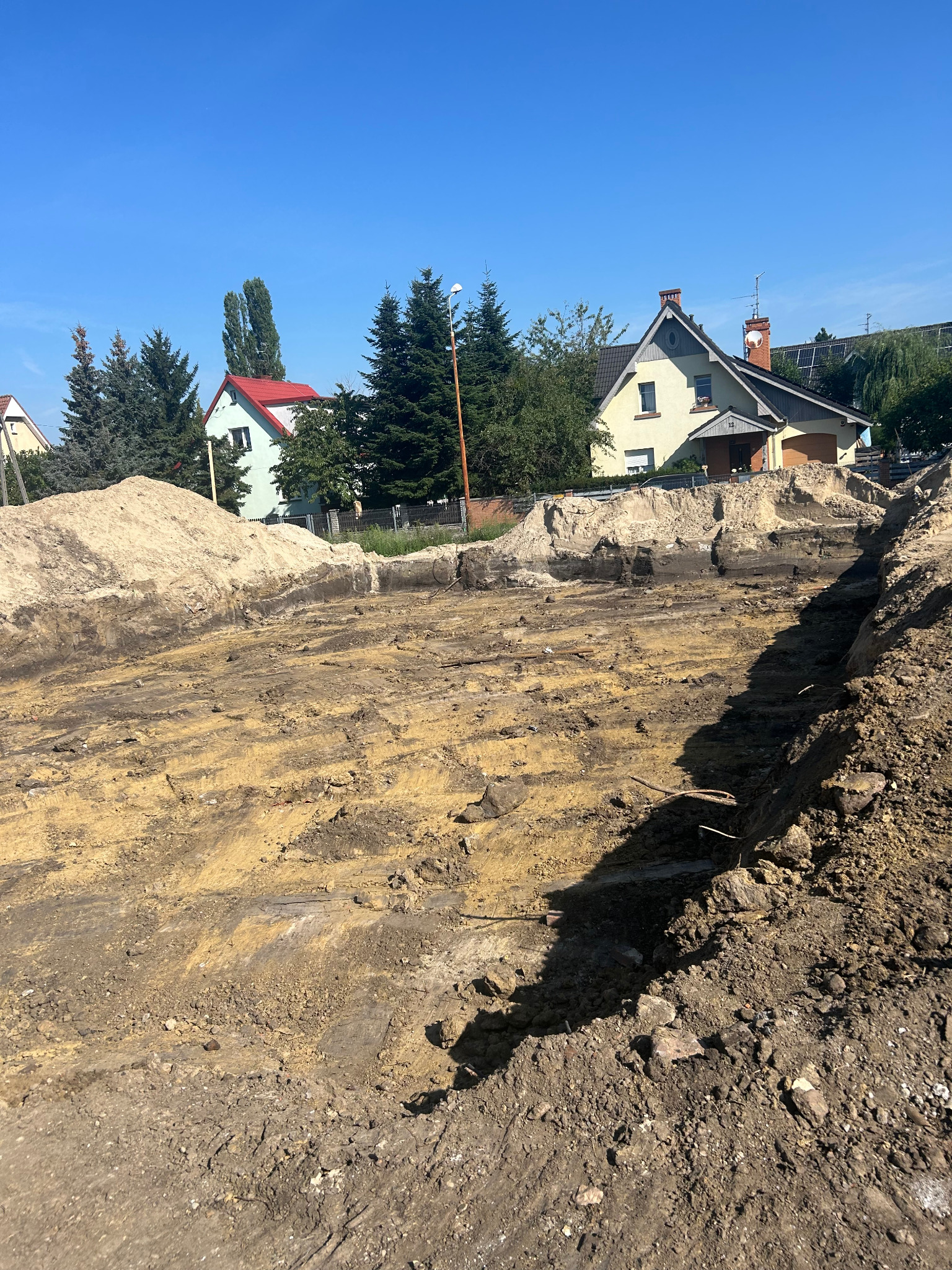 Wykop pod fundamenty domu jednorodzinnego w słoneczny dzień. Widoczne ślady po pracy koparki i hałdy ziemi. W tle zabudowa mieszkalna.