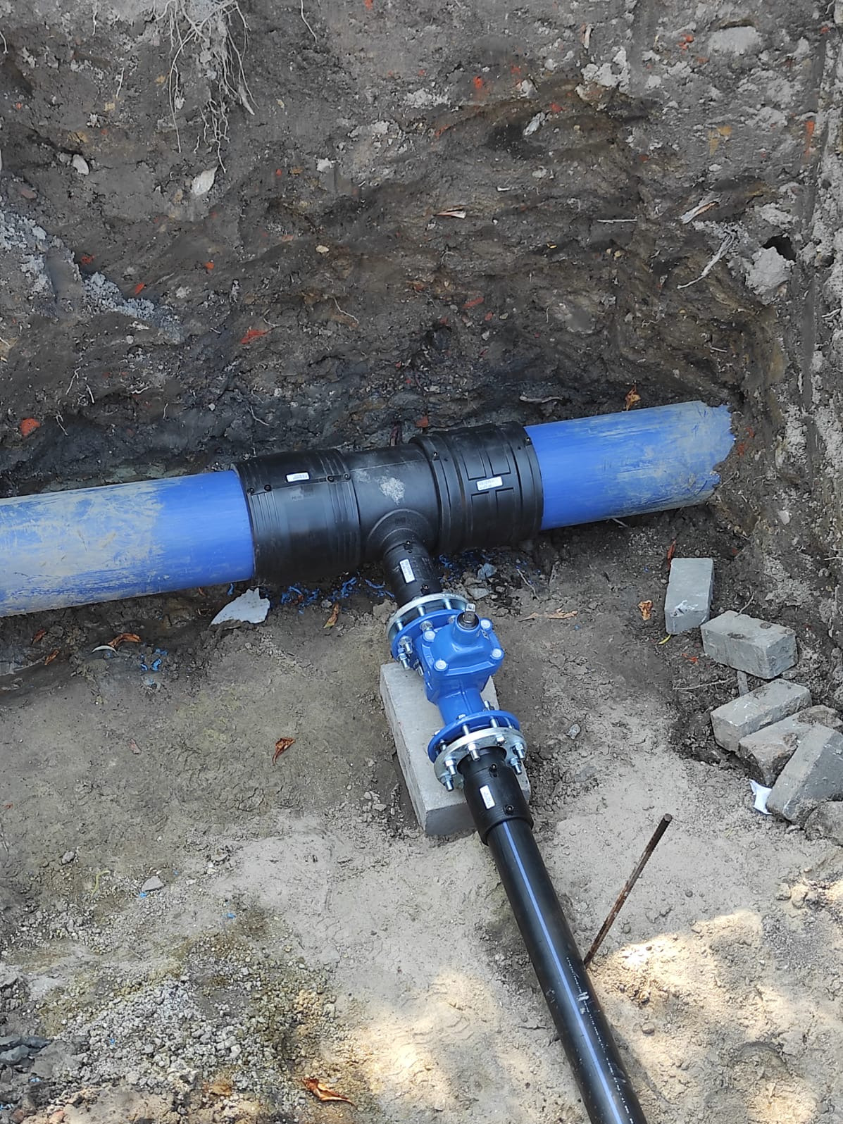 Połączenie niebieskiego rurociągu z zaworem w wykopie ziemnym. Widoczne złącza, rury z tworzywa i fragmenty gruzu na dnie wykopu. Instalacja wodociągowa w trakcie montażu.