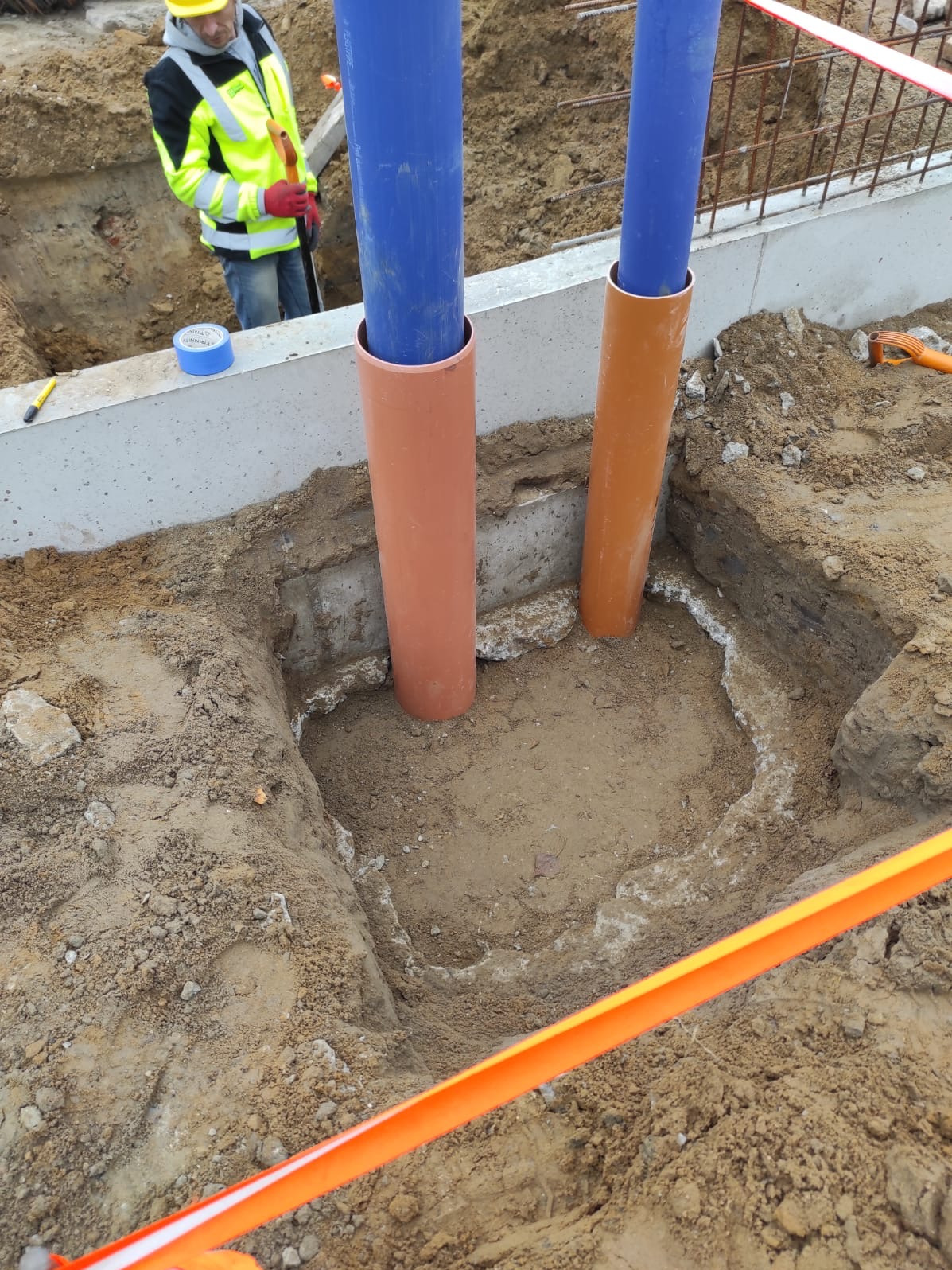 Wykop pod instalacje w gruncie we Wrocławiu, zabezpieczony pomarańczową taśmą. Widoczne wystające rury w osłonach. Pracownik w tle.