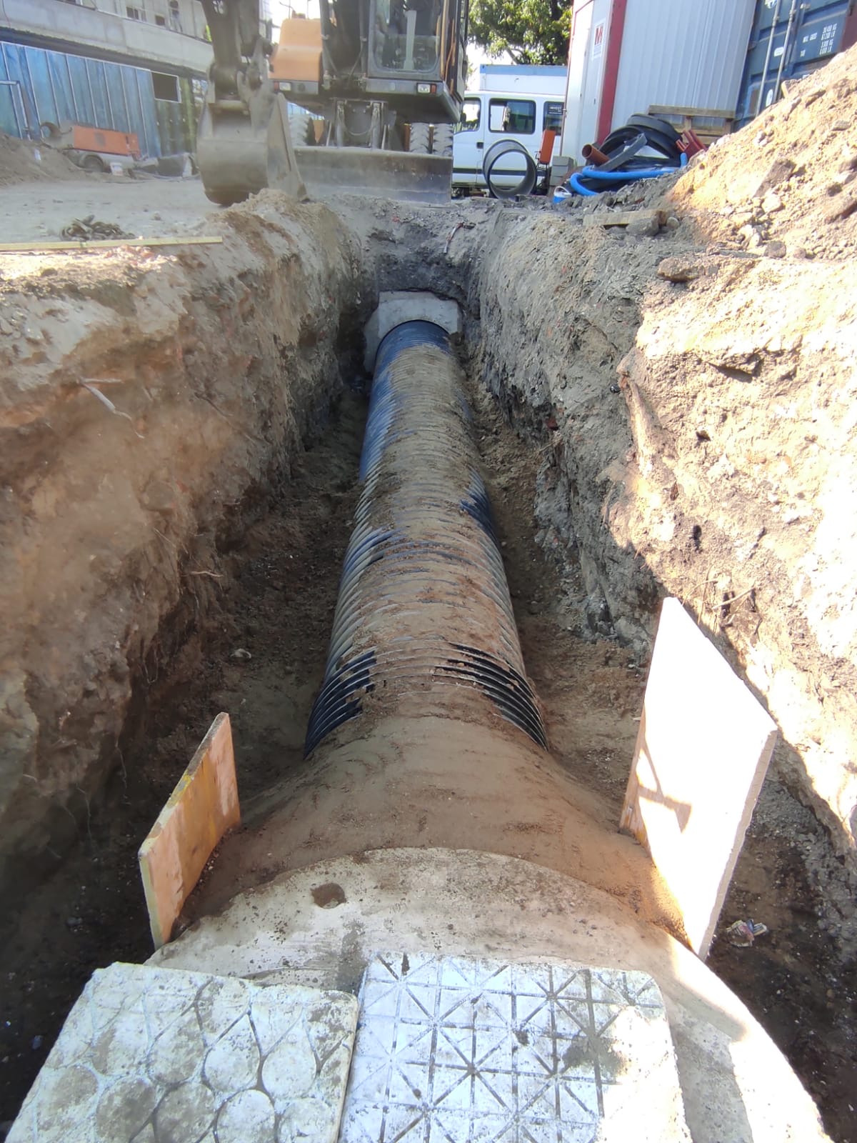 Długi, karbowany rurociąg w wykopie ziemnym z widocznym fragmentem koparki i pojazdami w tle. Ujęcie z perspektywy wnętrza wykopu.