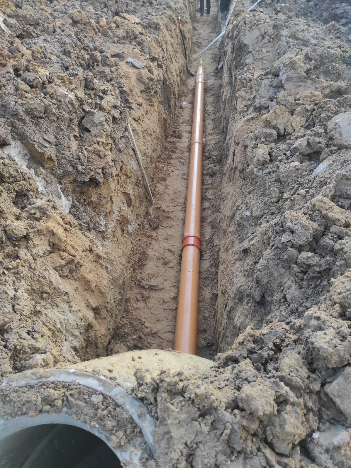 Brązowa rura kanalizacyjna z czerwonym złączem ułożona w wykopie ziemnym. Widoczne ślady pracy ziemnej i fragment betonowego kręgu.