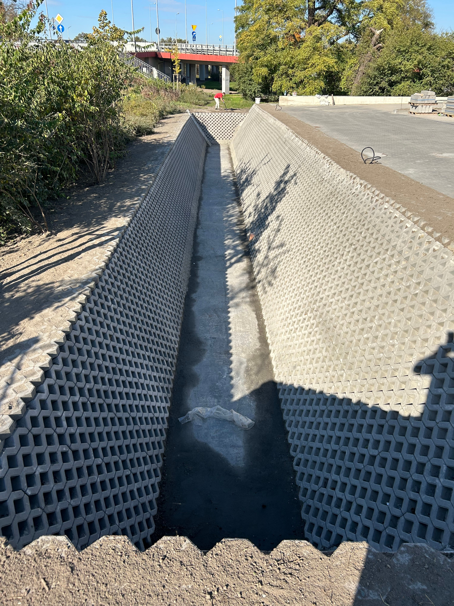 Długi wykop wyłożony ażurowymi betonowymi płytami, prawdopodobnie pod instalacje. Widoczny fragment terenu z zielenią i element infrastruktury drogowej w tle.
