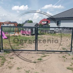 PRO Panel Ogrodzenia - Nowoczesna, grafitowa brama panelowa z furtką, osadzona na betonowych słupkach. W tle plac zabaw i dom jednorodzinny w słoneczny dzień.