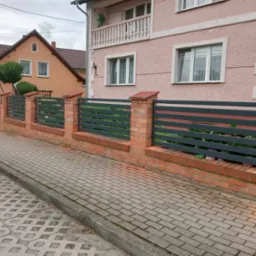 Nowoczesne ogrodzenie z poziomych, grafitowych paneli i ceglanych słupków, oddzielające posesję od chodnika z kostki. W tle różowy dom z balkonem.