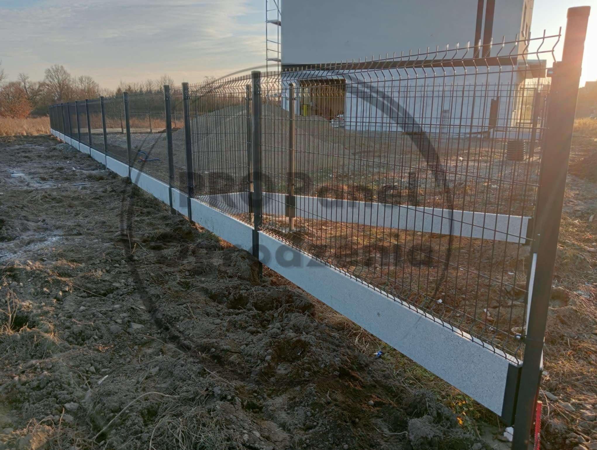 Panelowe ogrodzenie z podmurówką betonową na nierównym terenie, widok z perspektywy ziemi, w tle budynek i drzewa w pochmurny dzień.