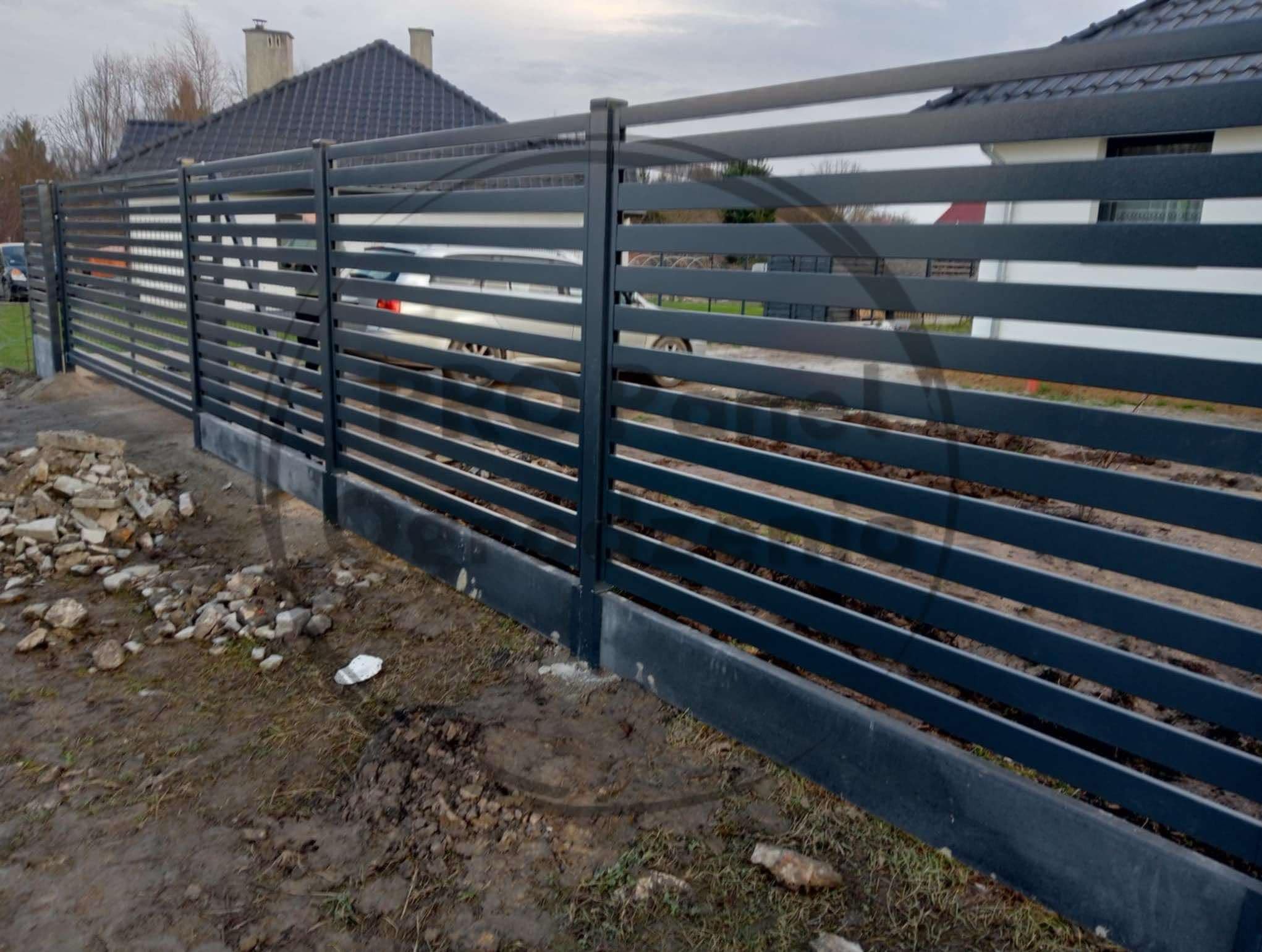 Nowoczesne, poziome ogrodzenie metalowe w kolorze grafitowym, z widocznymi elementami betonowej podmurówki i fragmentem posesji w tle. Widoczne ślady prac ziemnych.