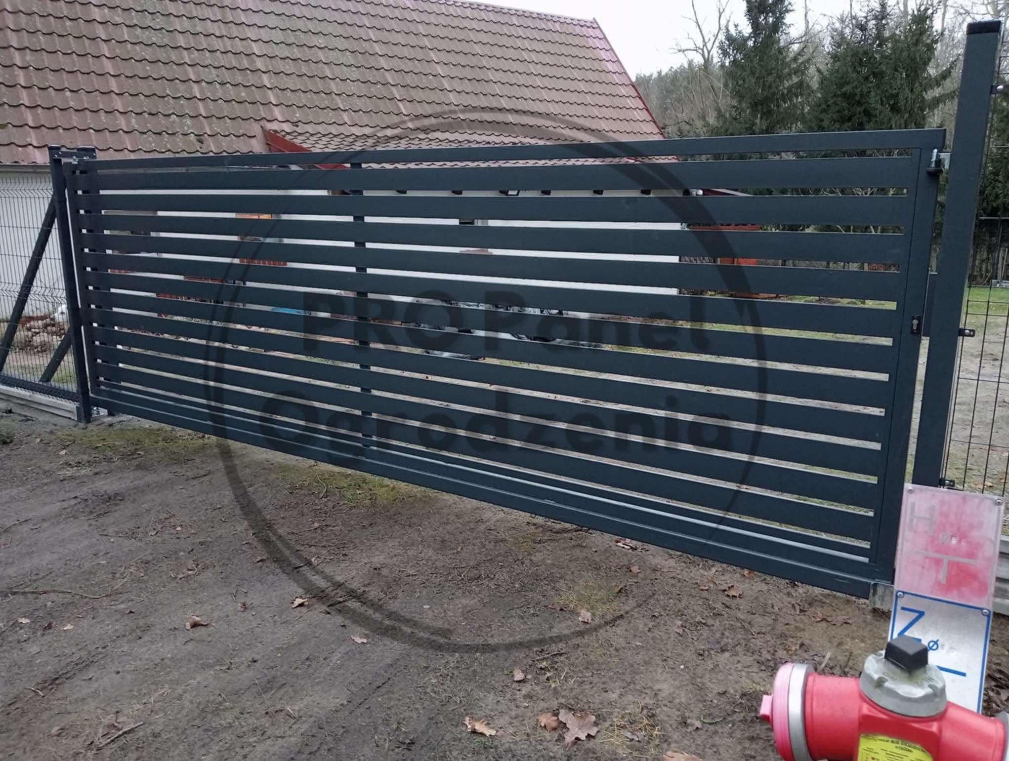 Nowoczesna, przesuwna brama metalowa w kolorze grafitowym z poziomymi szczeblami, na tle domu z czerwoną dachówką i zieleni. Widoczny hydrant.