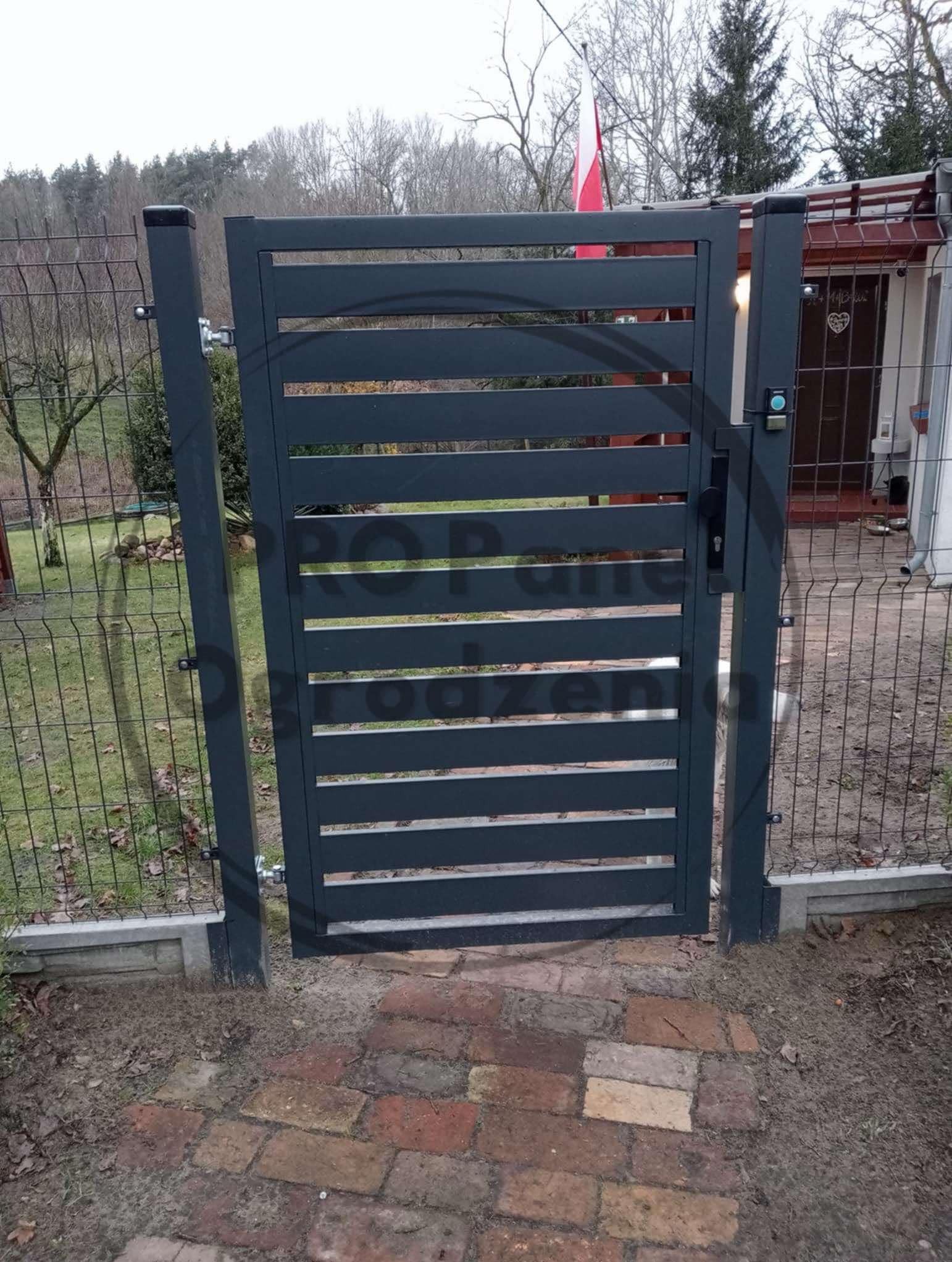 Nowoczesna, metalowa brama posesyjna z poziomymi szczeblami, zamontowana przy ogrodzeniu z siatki. Na posesji powiewa polska flaga. Czerwona cegła na ścieżce.