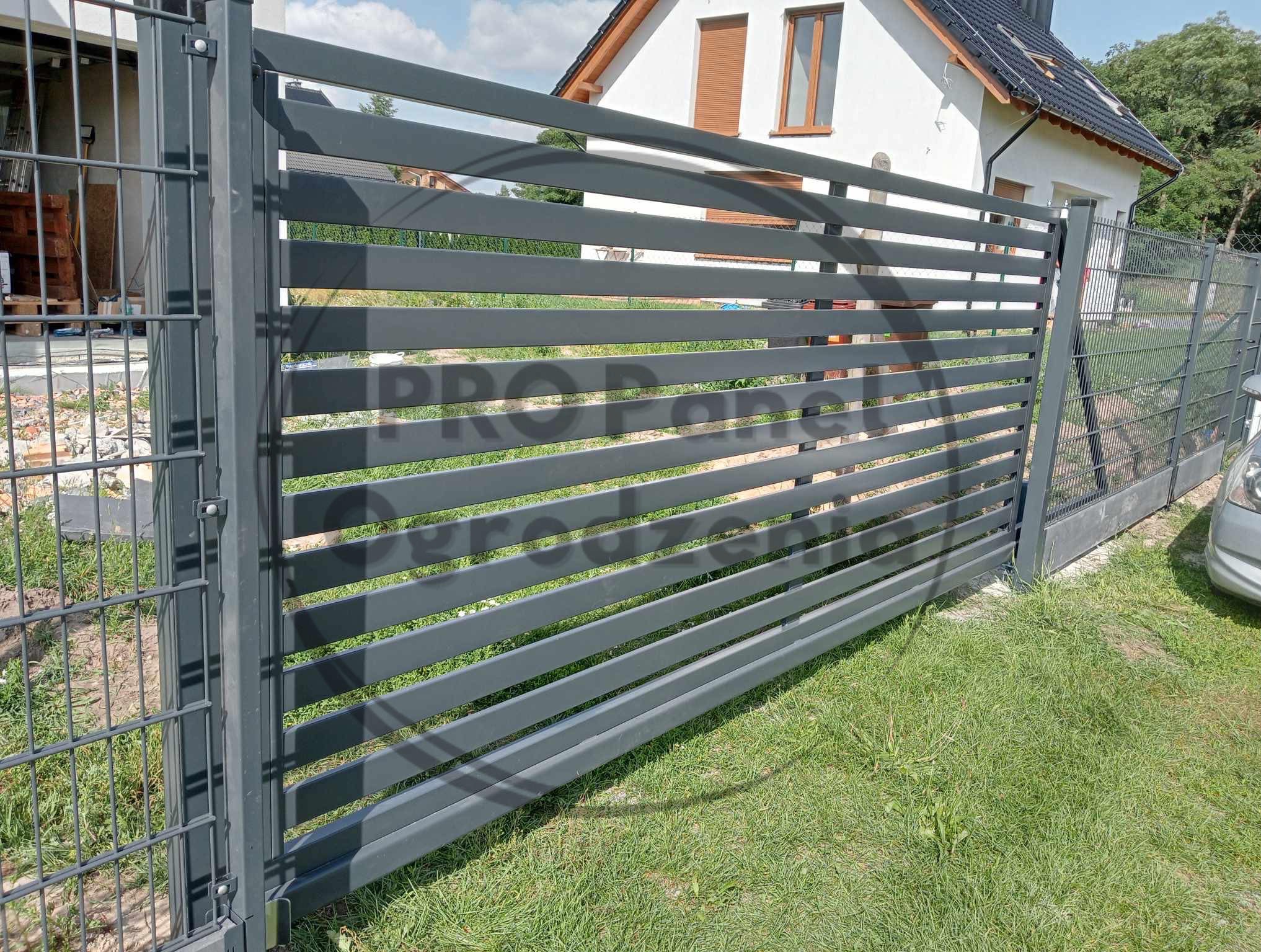 Nowoczesna, pozioma brama panelowa w kolorze grafitowym, z widocznym fragmentem domu w tle, na tle zielonego trawnika. Brama zamontowana.