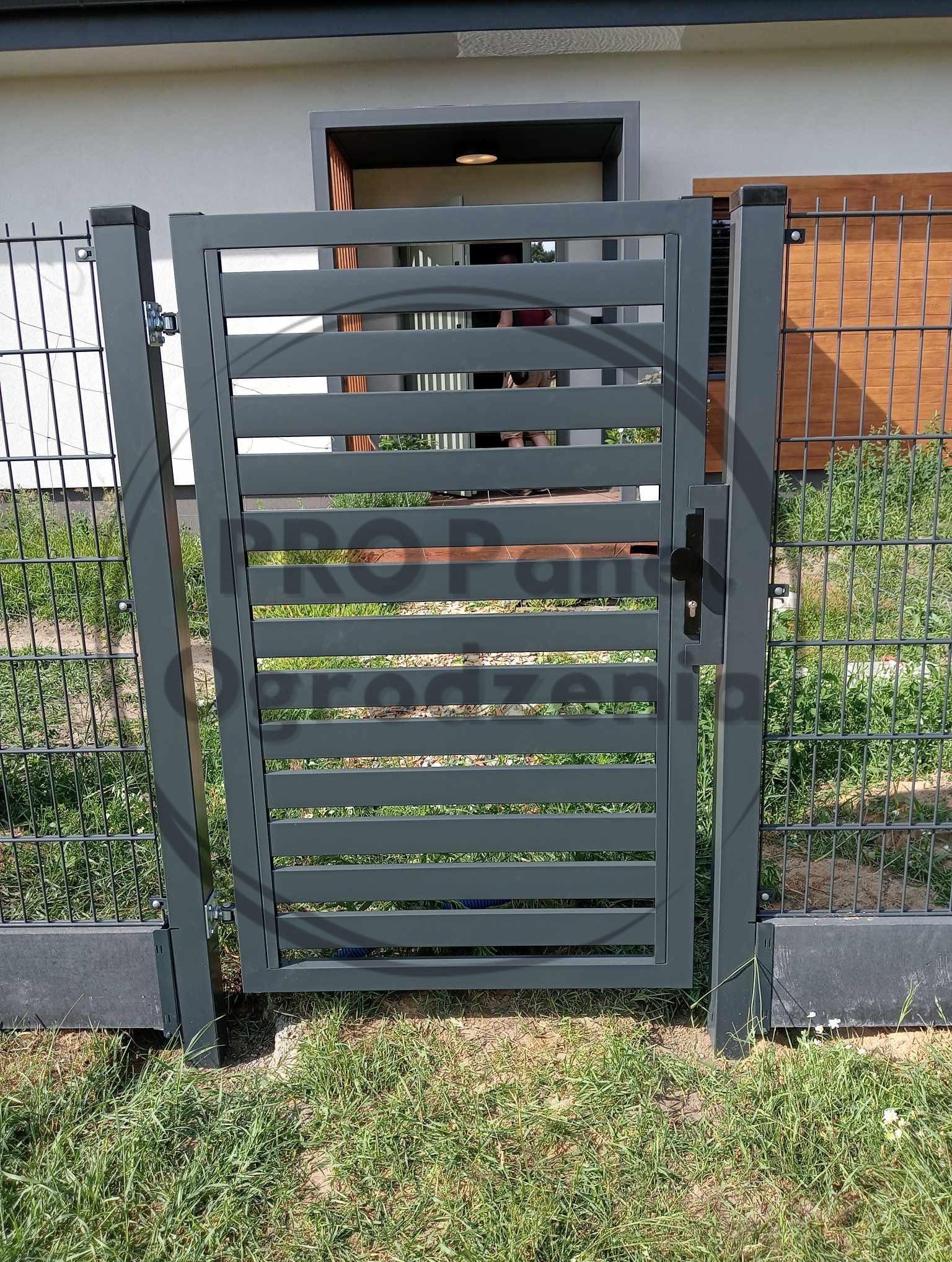 Nowoczesna, szara, metalowa furtka z poziomymi szczeblami, wkomponowana w ogrodzenie panelowe. Widoczny zamek i klamka. Trawa w tle.