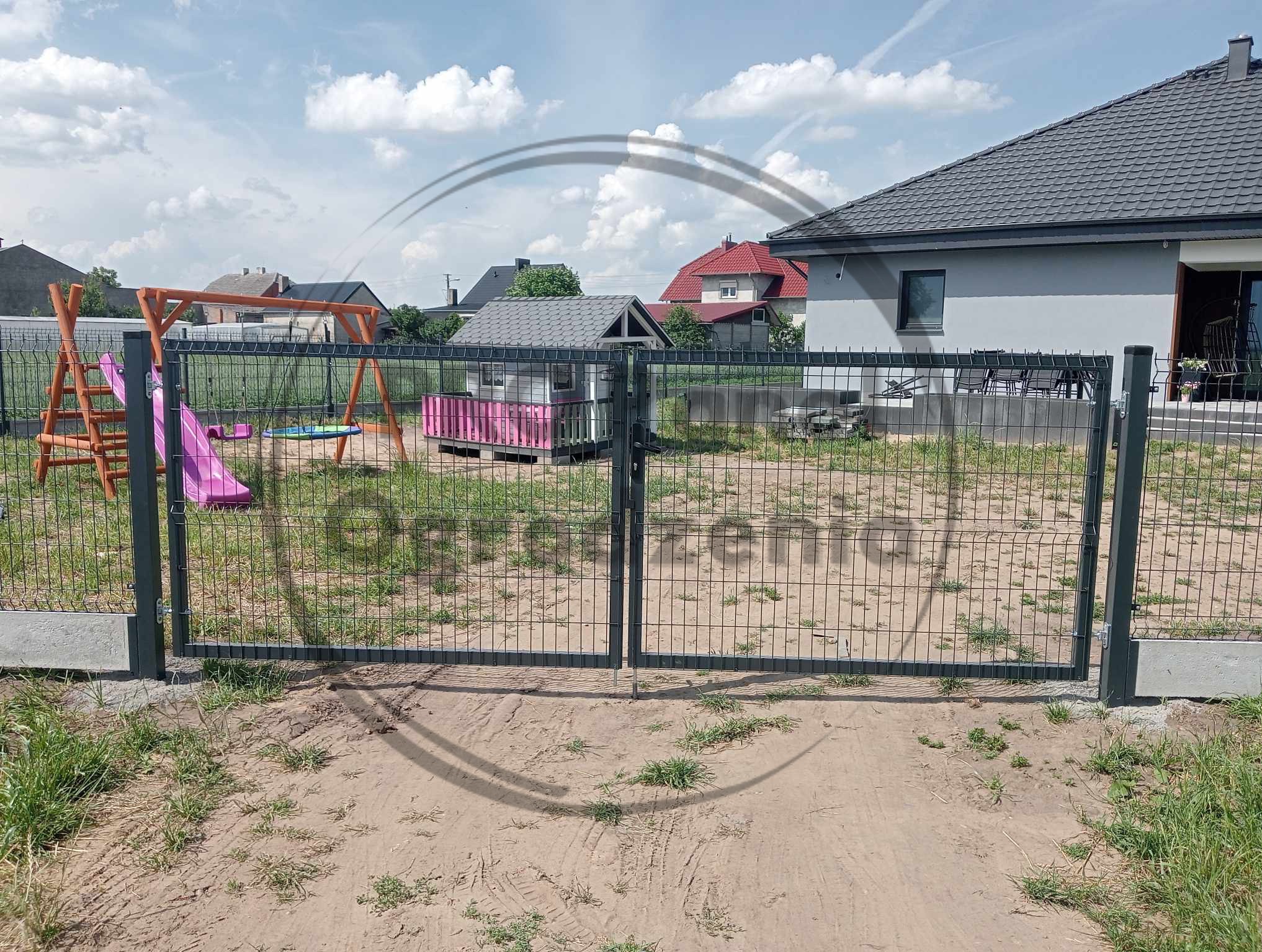 Nowoczesna, grafitowa brama panelowa z furtką, osadzona na betonowych słupkach. W tle plac zabaw i dom jednorodzinny w słoneczny dzień.