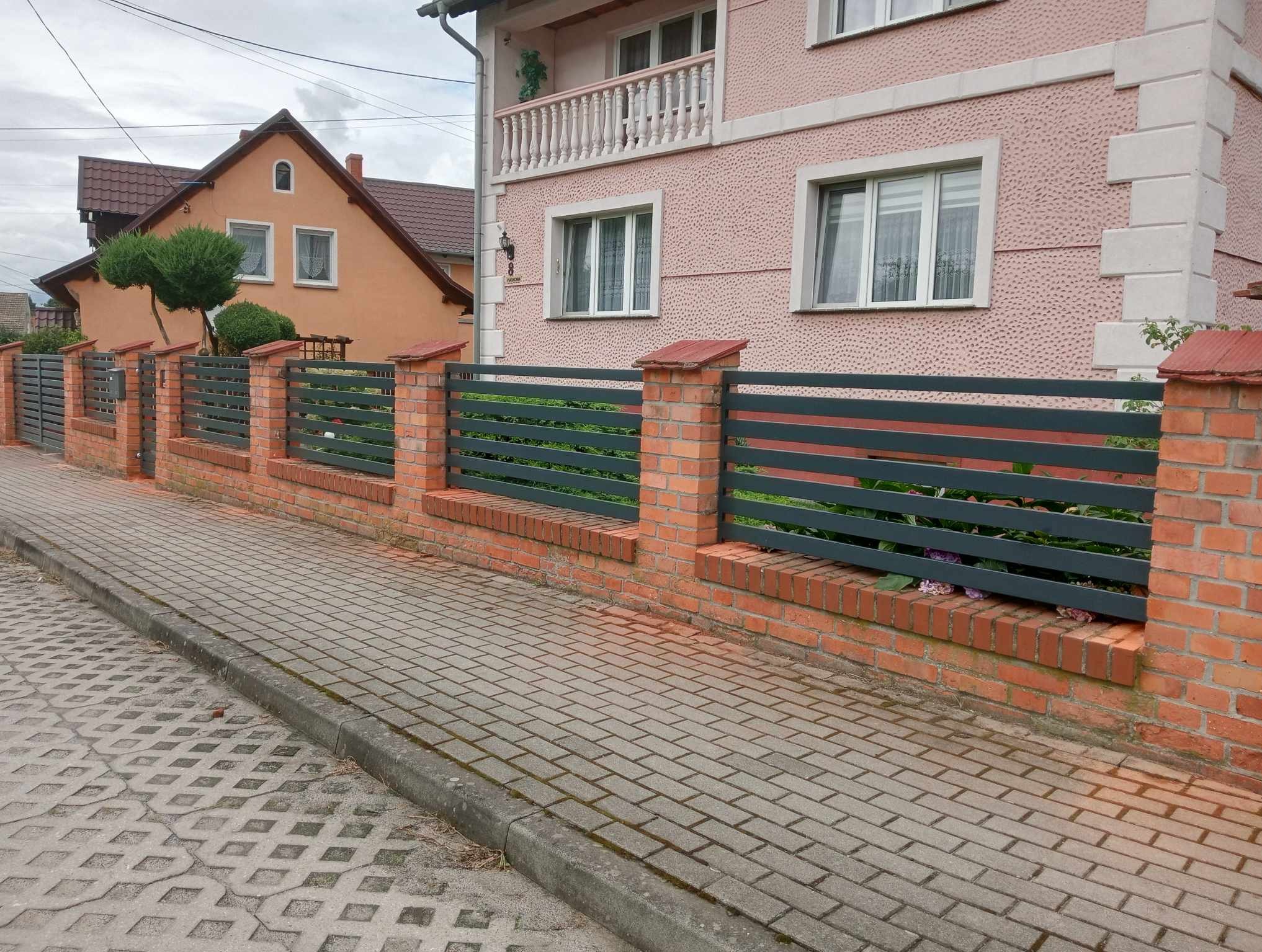 Nowoczesne ogrodzenie z poziomych, grafitowych paneli i ceglanych słupków, oddzielające posesję od chodnika z kostki. W tle różowy dom z balkonem.