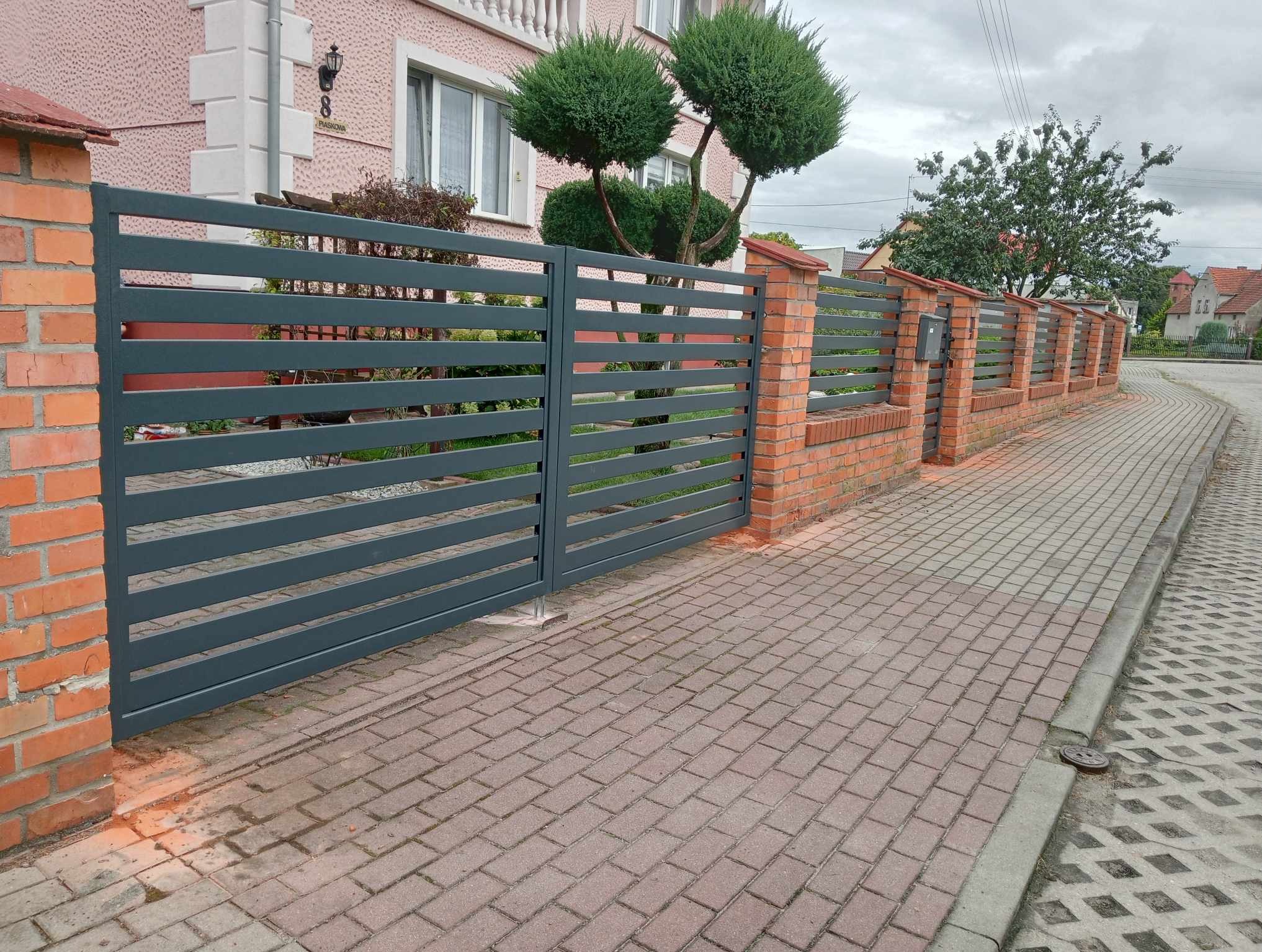 Nowoczesna brama wjazdowa i ogrodzenie panelowe z poziomych, szarych profili, osadzone na ceglanych słupkach. Widok z zewnątrz posesji, kostka brukowa.