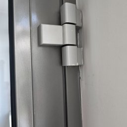 Technanika Sebastian Olszowski - Szare drzwi z widocznym, białym zawiasem przymocowanym do ściany. Detal ukazuje precyzję montażu i solidność konstrukcji.