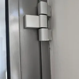 Szare drzwi z widocznym, białym zawiasem przymocowanym do ściany. Detal ukazuje precyzję montażu i solidność konstrukcji.