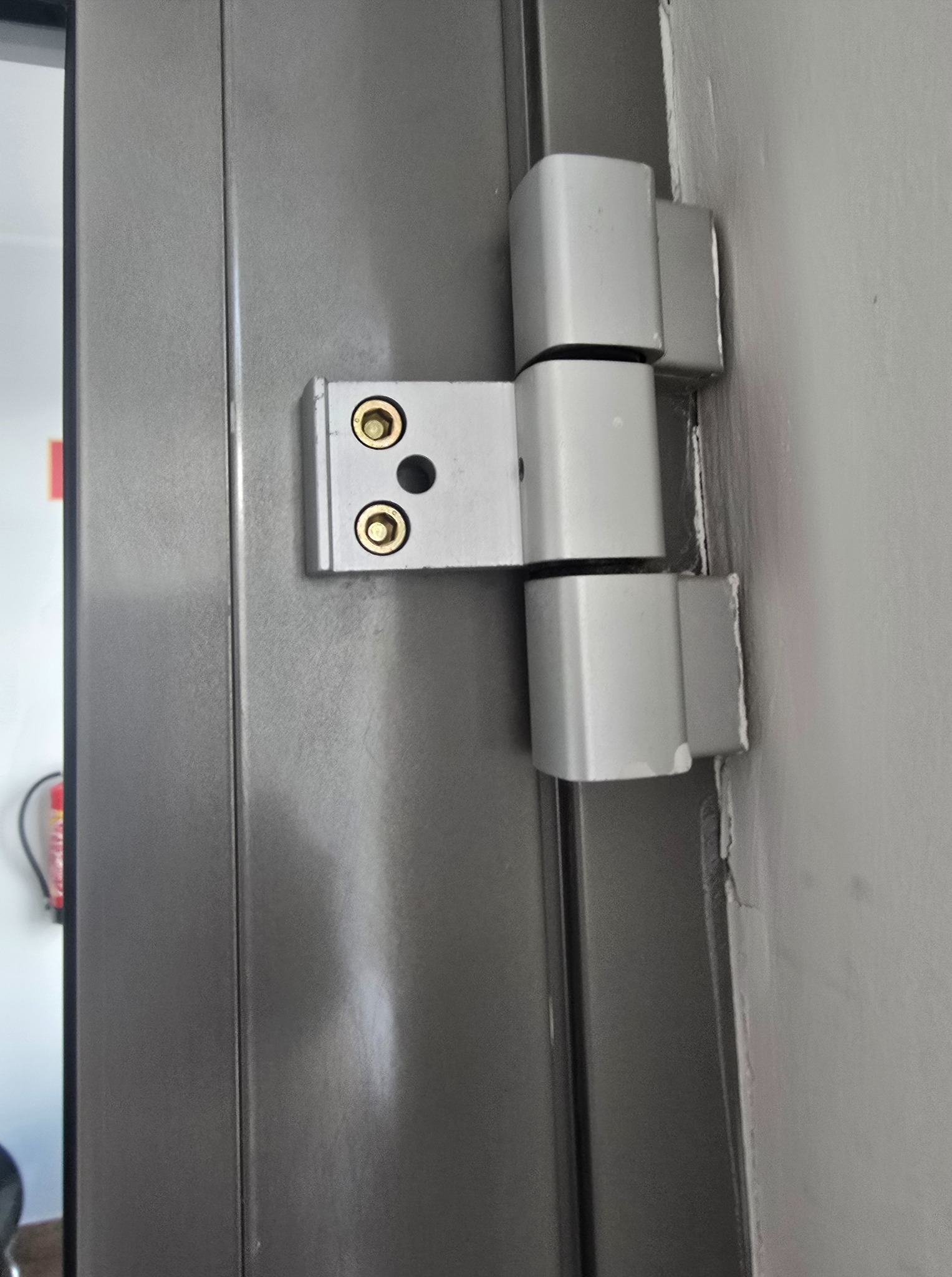 Szary zawias drzwiowy z widocznymi śrubami mocującymi, zamontowany na szarej futrynie i ścianie. Widoczne ślady użytkowania na ścianie przy zawiasie.