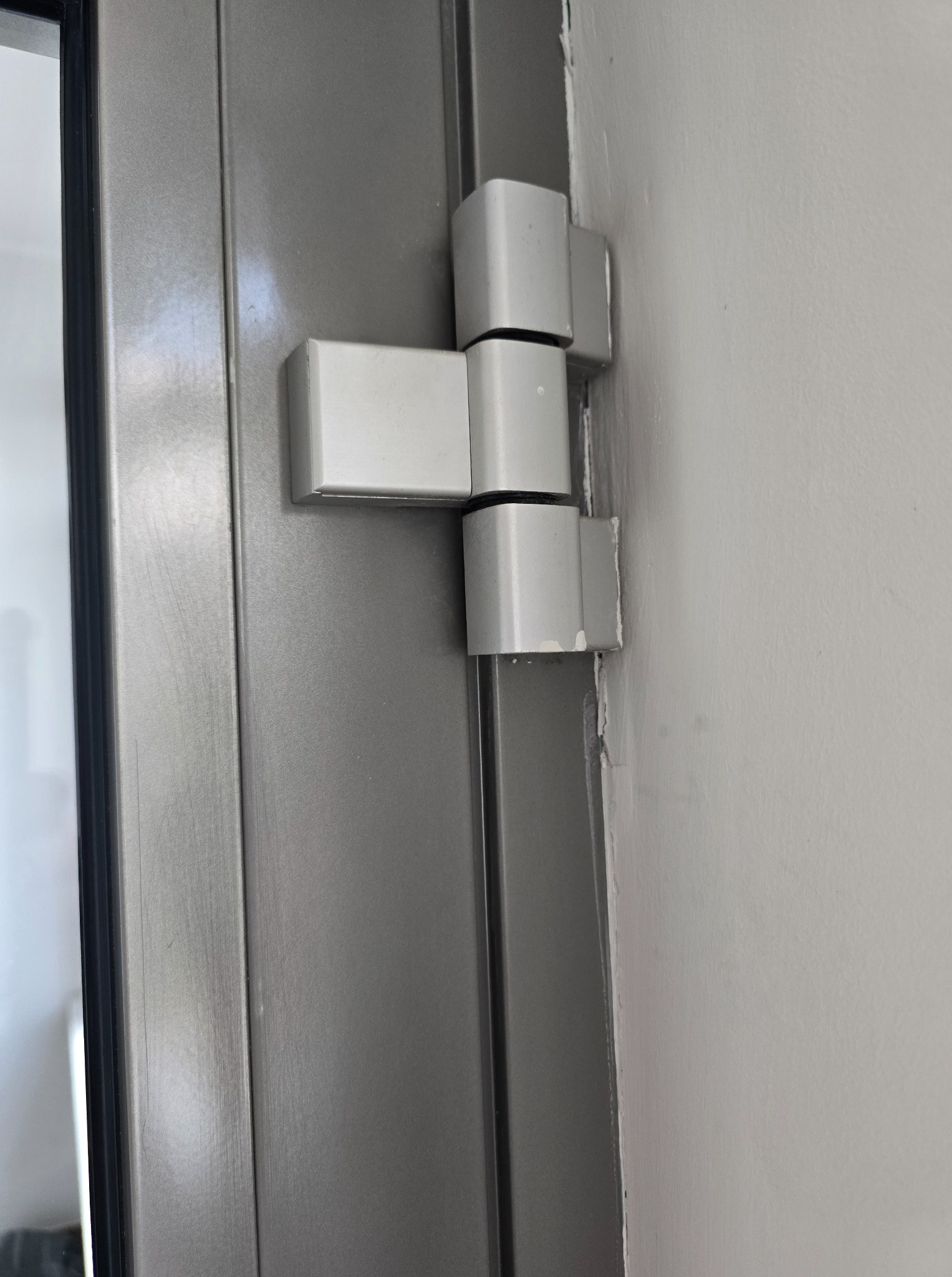 Szare drzwi z widocznym, białym zawiasem przymocowanym do ściany. Detal ukazuje precyzję montażu i solidność konstrukcji.