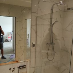 4L4 - Nowoczesna kabina prysznicowa z marmurowymi płytkami, szklaną ścianką i deszczownicą. Widoczne przygotowania do montażu armatury i odbicie osoby w lustrze.