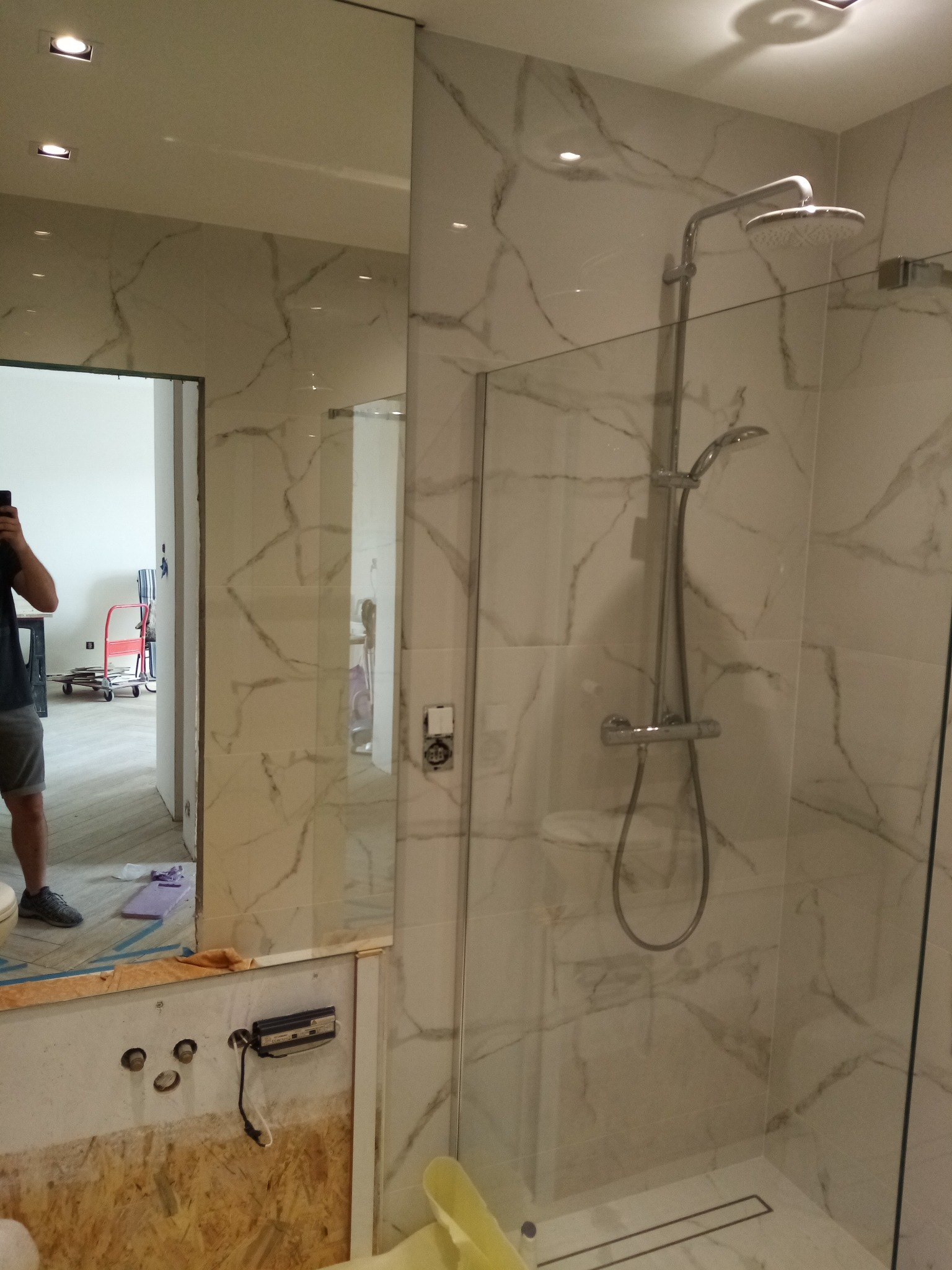 Nowoczesna kabina prysznicowa z marmurowymi płytkami, szklaną ścianką i deszczownicą. Widoczne przygotowania do montażu armatury i odbicie osoby w lustrze.