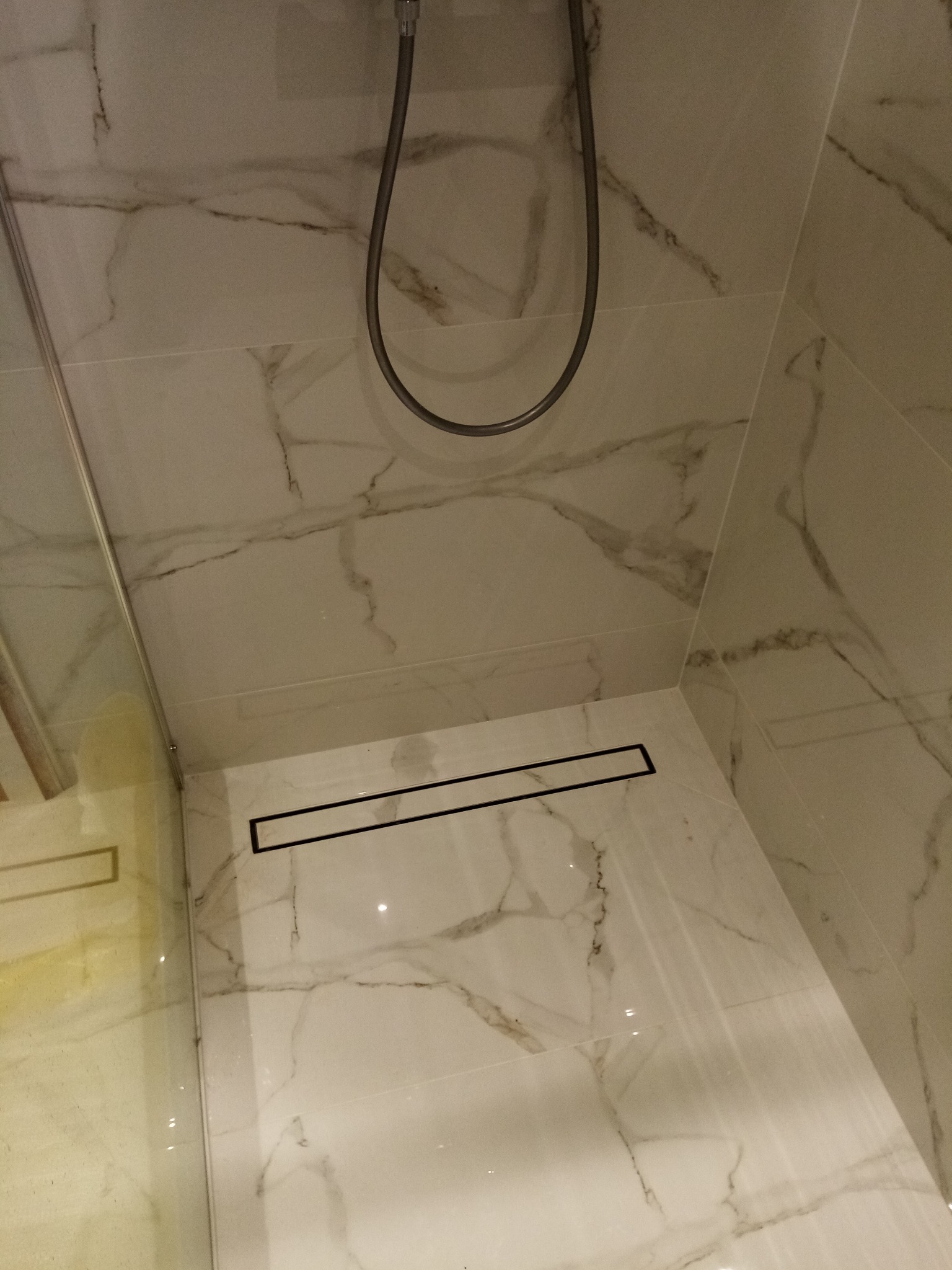 Nowoczesny odpływ liniowy w kabinie prysznicowej wyłożonej białymi płytkami imitującymi marmur z delikatnym, szarym żyłkowaniem. Widoczna słuchawka prysznicowa.