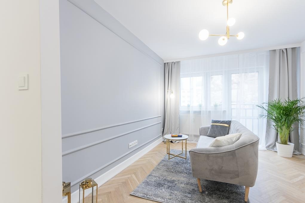 Elegancki salon w odcieniach szarości: sofa, stolik kawowy na złotych nóżkach, parkiet w jodełkę, sztukateria na ścianie i roślina doniczkowa przy oknie.