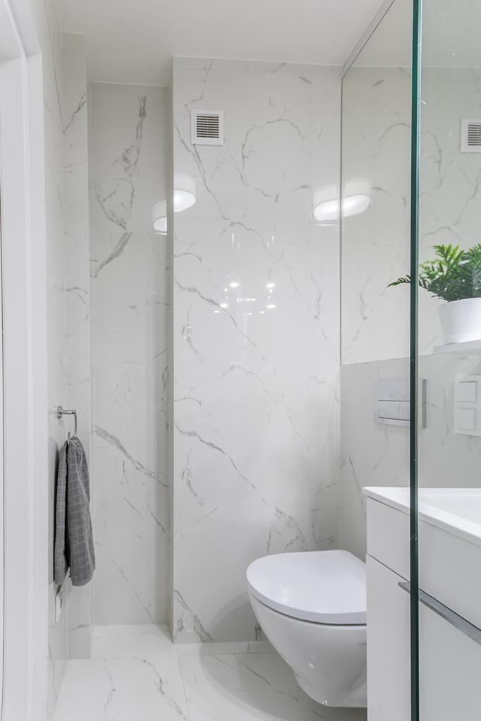 Jasna łazienka z białymi płytkami imitującymi marmur, wisząca toaleta, szklana kabina prysznicowa i szary ręcznik na wieszaku. Minimalistyczny design.